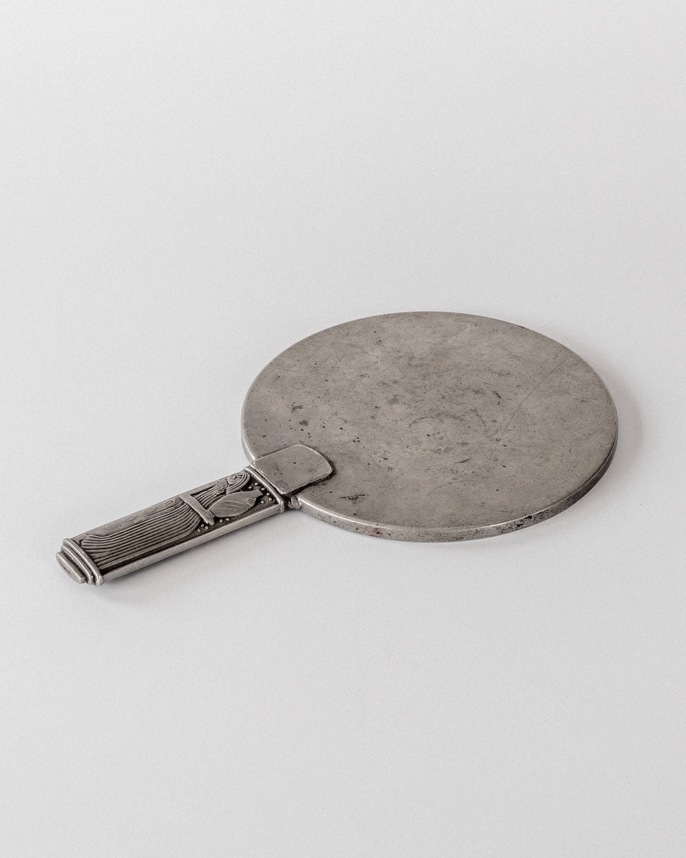 A vintage round silver pewter hand mirror with a geometric-patterned handle, placed on a white surface. Echoing Sylvia Stave's 1930s modernist design, the "Modernist Metal Mirror" by Spigel appears tarnished yet elegant.