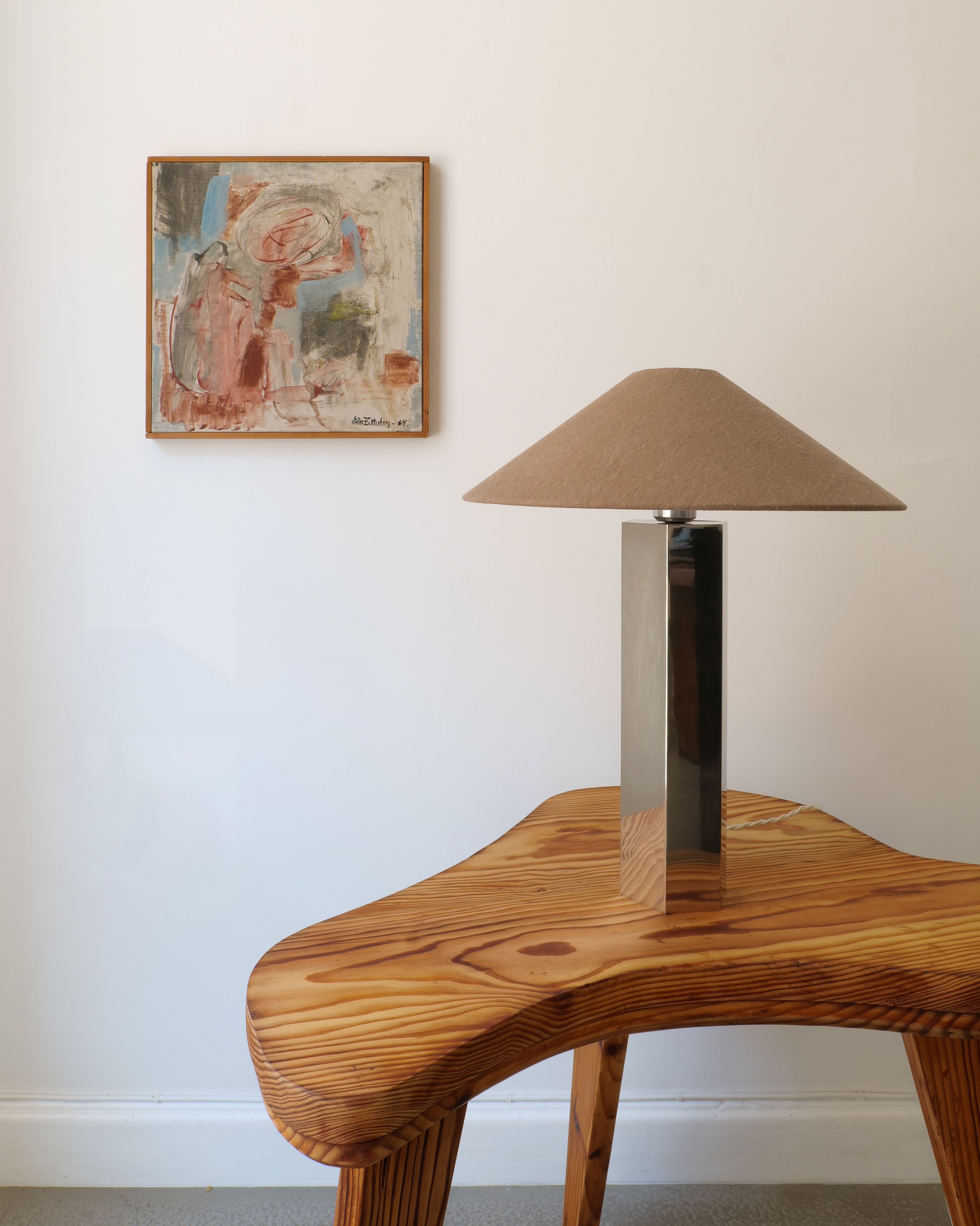 A modern wooden side table with a sleek lamp featuring a rectangular metal base and beige conical shade stands in the foreground, with 'Summer', a 1960s painting by Swedish artist Ester Zetterberg from Collection apart, adorning the white wall behind it.