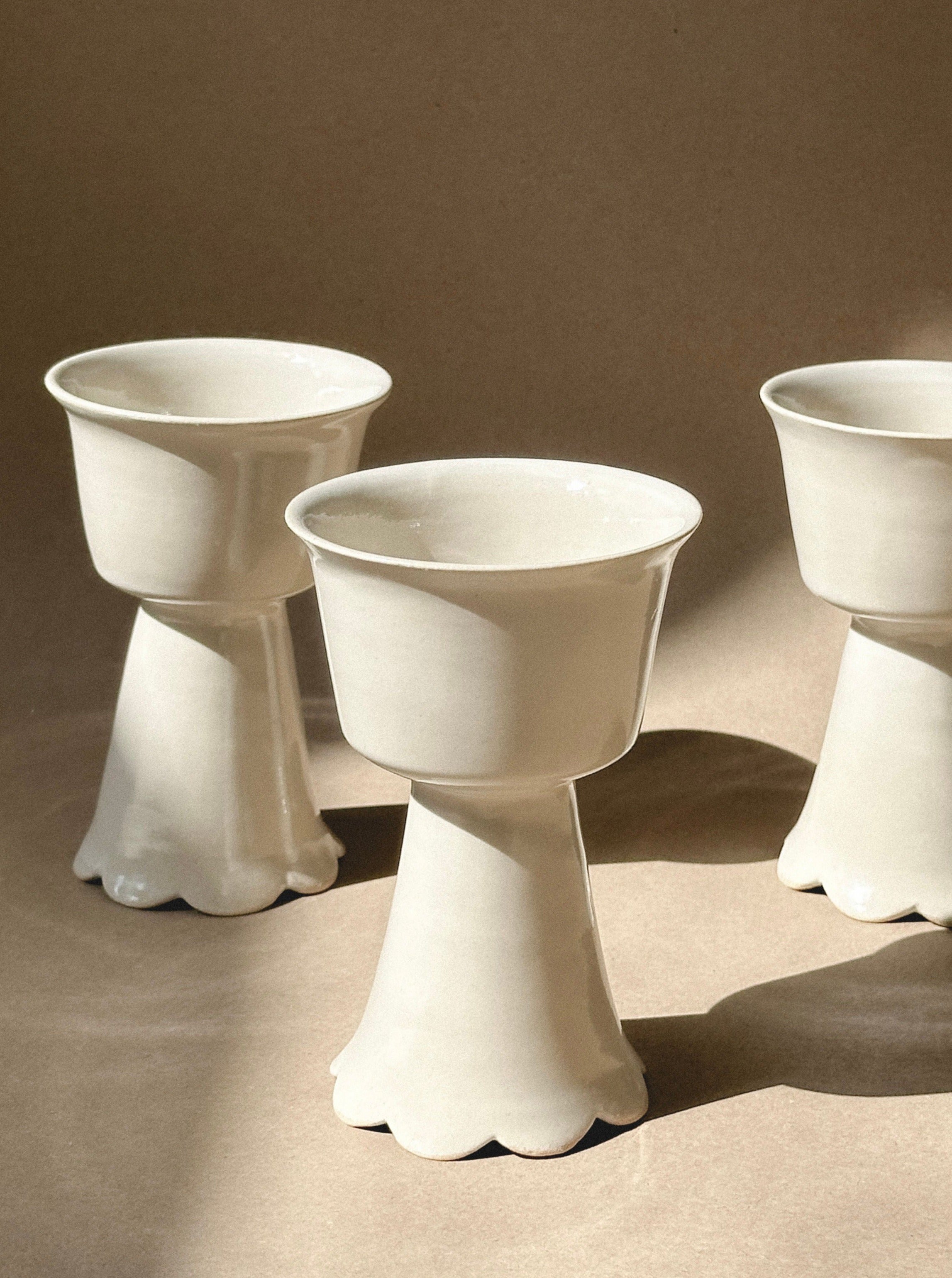 Three Flowery Wine Goblets by noki ceramics with scalloped bases, reminiscent of handmade stoneware, are arranged on a beige surface. The goblets have a glossy finish, and sunlight highlights their smooth texture, casting soft shadows.