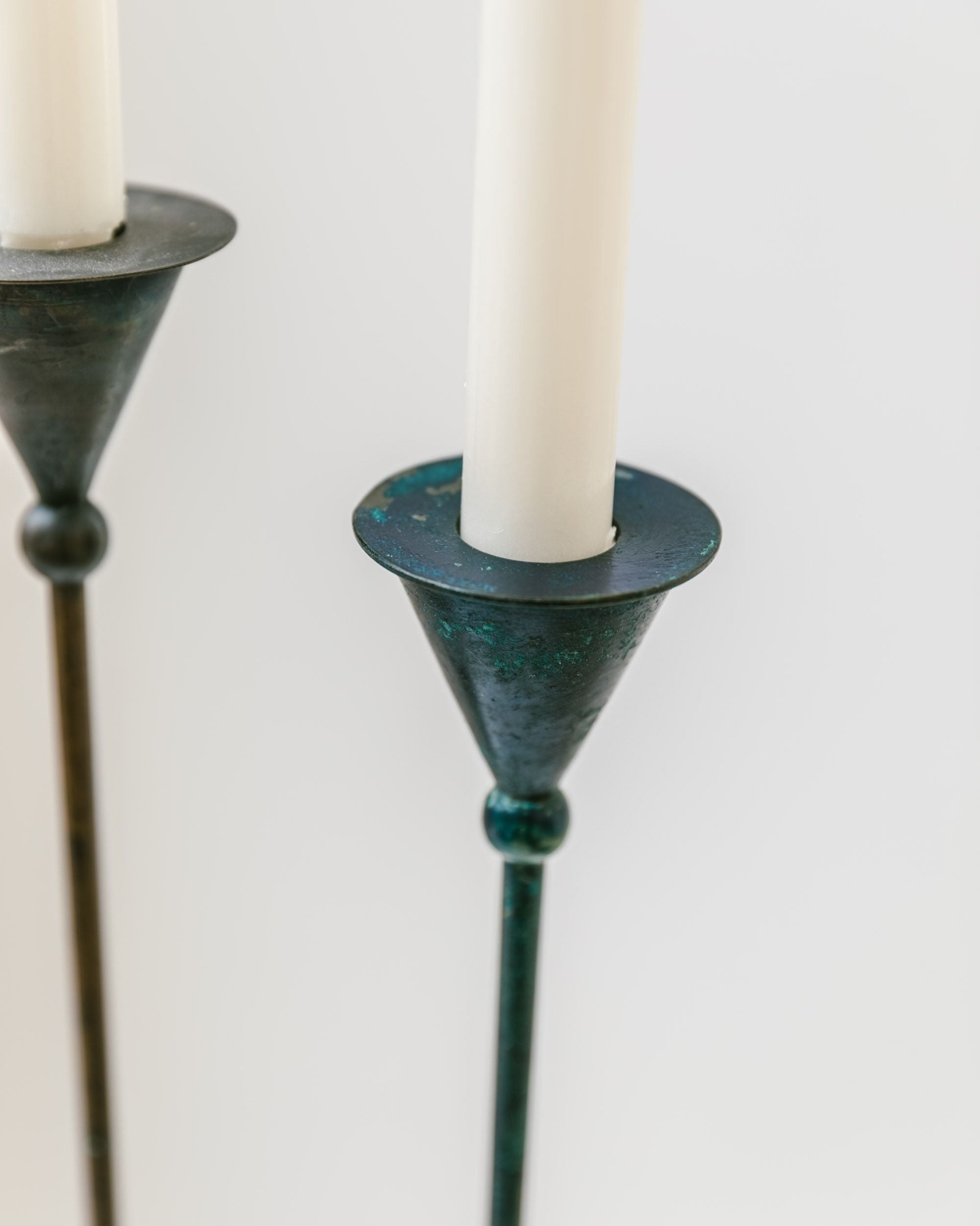 Close-up of a set of two tall, slender candlesticks from Bottega Jacobs, featuring white candles with a conical form. These Postmodern 1980s vintage candleholders boast dark, weathered metal bases with a patina, elegantly displayed against a plain, light background.