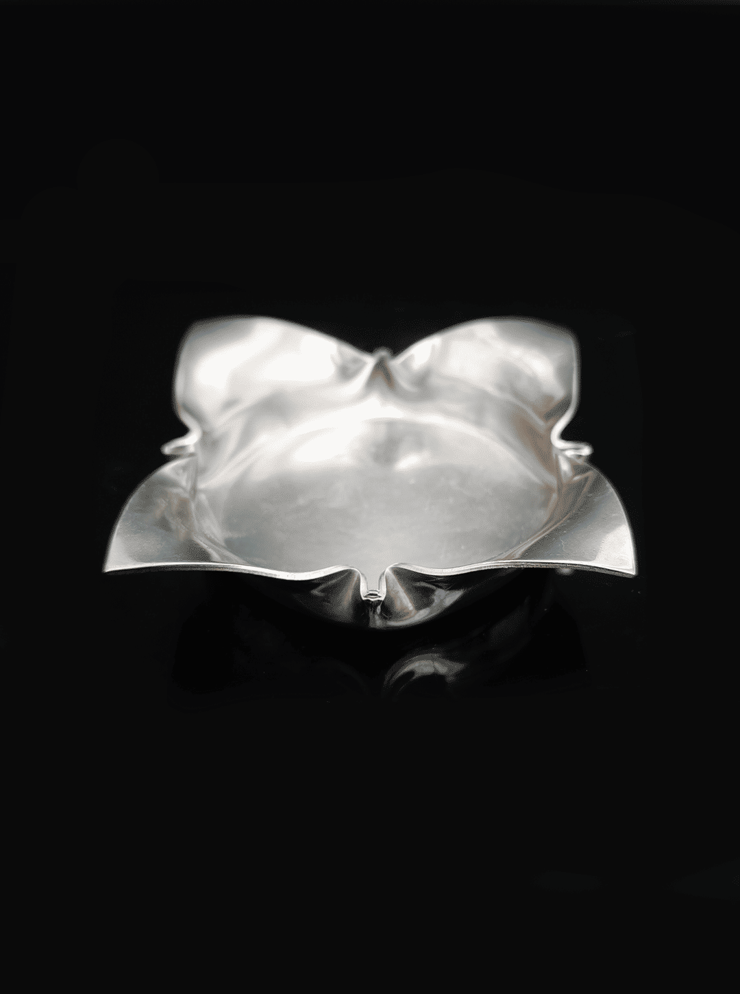 The Les Objoies Folded Ashtray is a silver, flower-shaped dish with petal-like edges and a smooth surface, set against a black background.