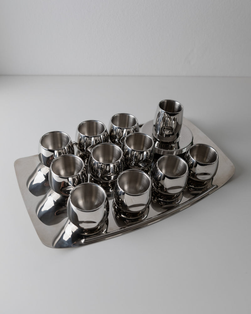 The Coffee set by AMC 70s from Bottega Jacobs features a minimalist rectangular metal tray with twelve small shiny metal cups neatly arranged on a white surface.