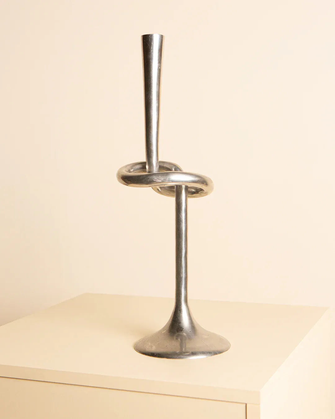 An Aluminum "Nœud" Candleholder 80's from Treaptyque, exemplifying modern elegance, rests on a light wooden surface. Its base gracefully curves into a knot before rising vertically, with a plain, light-colored wall serving as the background.