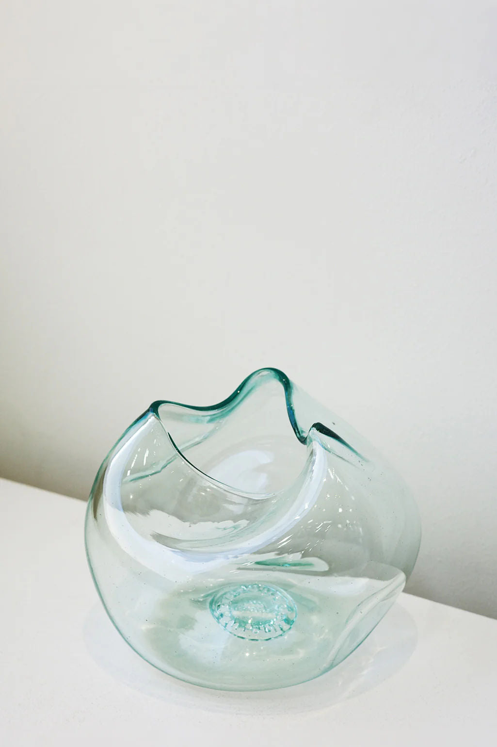 'The Bubble to End all Bubbles' by Completedworks, a transparent, hand-blown recycled glass vase in clear, with smooth curves and a wavy, leaf-like shape, sits on a white surface. It catches the light and creates subtle reflections against the plain background.