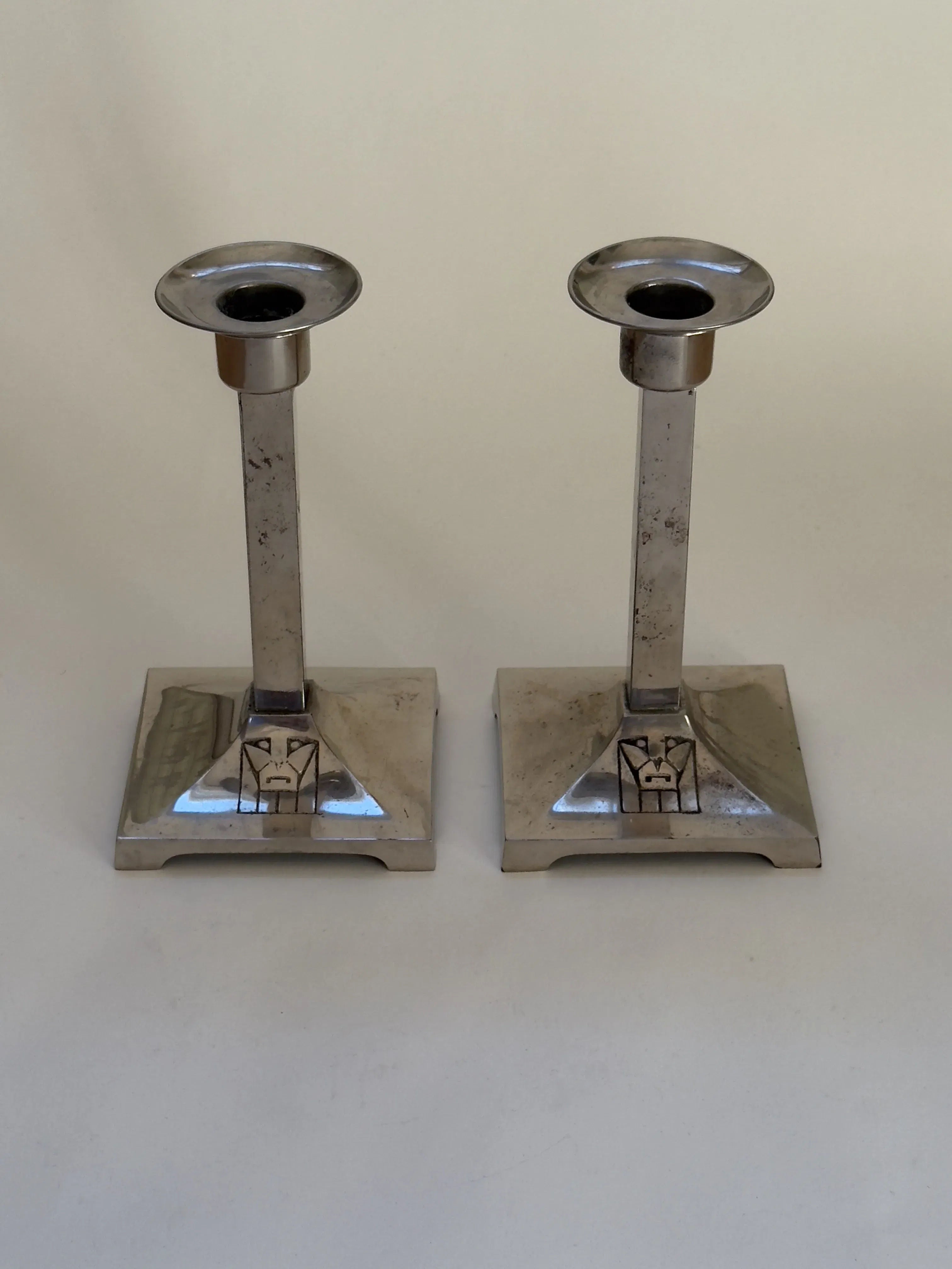 A pair of Galerie Storm's Art Deco candlesticks from the 20s/30s features geometric silhouettes and square bases on a neutral background. Each base has an engraved design near the stem, echoing early modernist style. The silver metal surfaces are slightly tarnished.