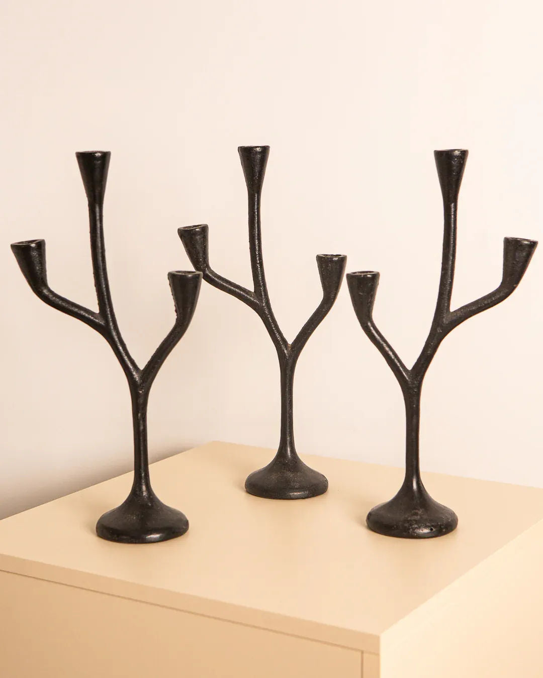 Three black cast aluminium 'Arbre' candleholders by Treaptyque, featuring an abstract branch-like design with multiple holders, showcase exquisite craftsmanship. Displayed on a light wooden surface against a beige wall, they offer a modern, artistic look.