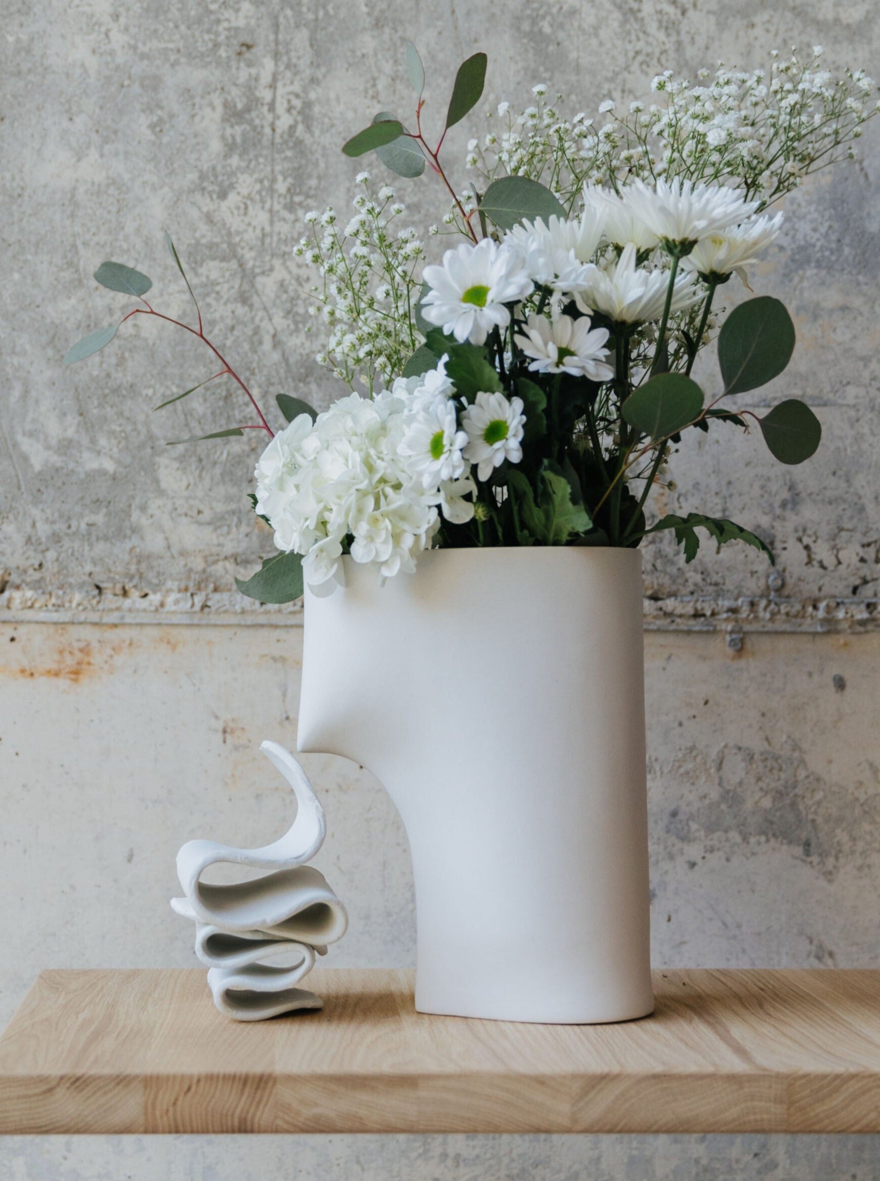 A one-of-a-kind Lisa Allegra Draped Vase Apparel, resembling a minimalist human profile, is adorned with white flowers, green leaves, and delicate baby's breath as it rests on a wooden surface. Next to it stands an abstract sculptural piece flaunting fluid forms in white porcelain. The backdrop features a textured, worn concrete wall.