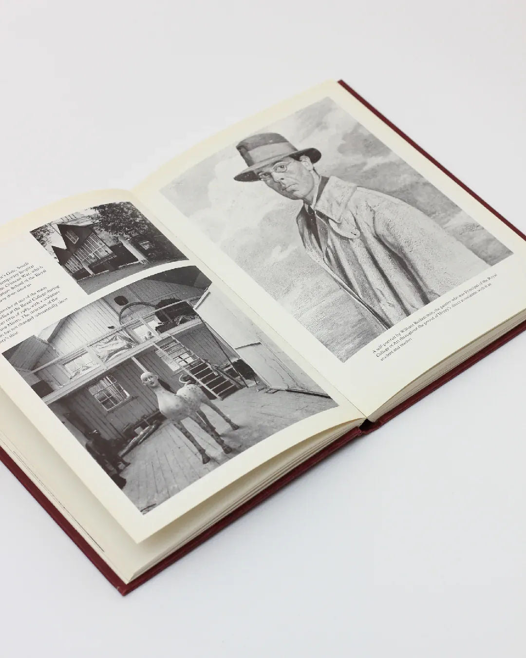 An open copy of "Henry Moore, An Illustrated Biography" by Boga Avante Shop showcases vintage black and white photographs. On the left page, there is an image of a man standing by a house; on the right page, there is a portrait of a man wearing a hat. The book has a red cover and rests on a white surface, echoing Henry Moore's timeless elegance.