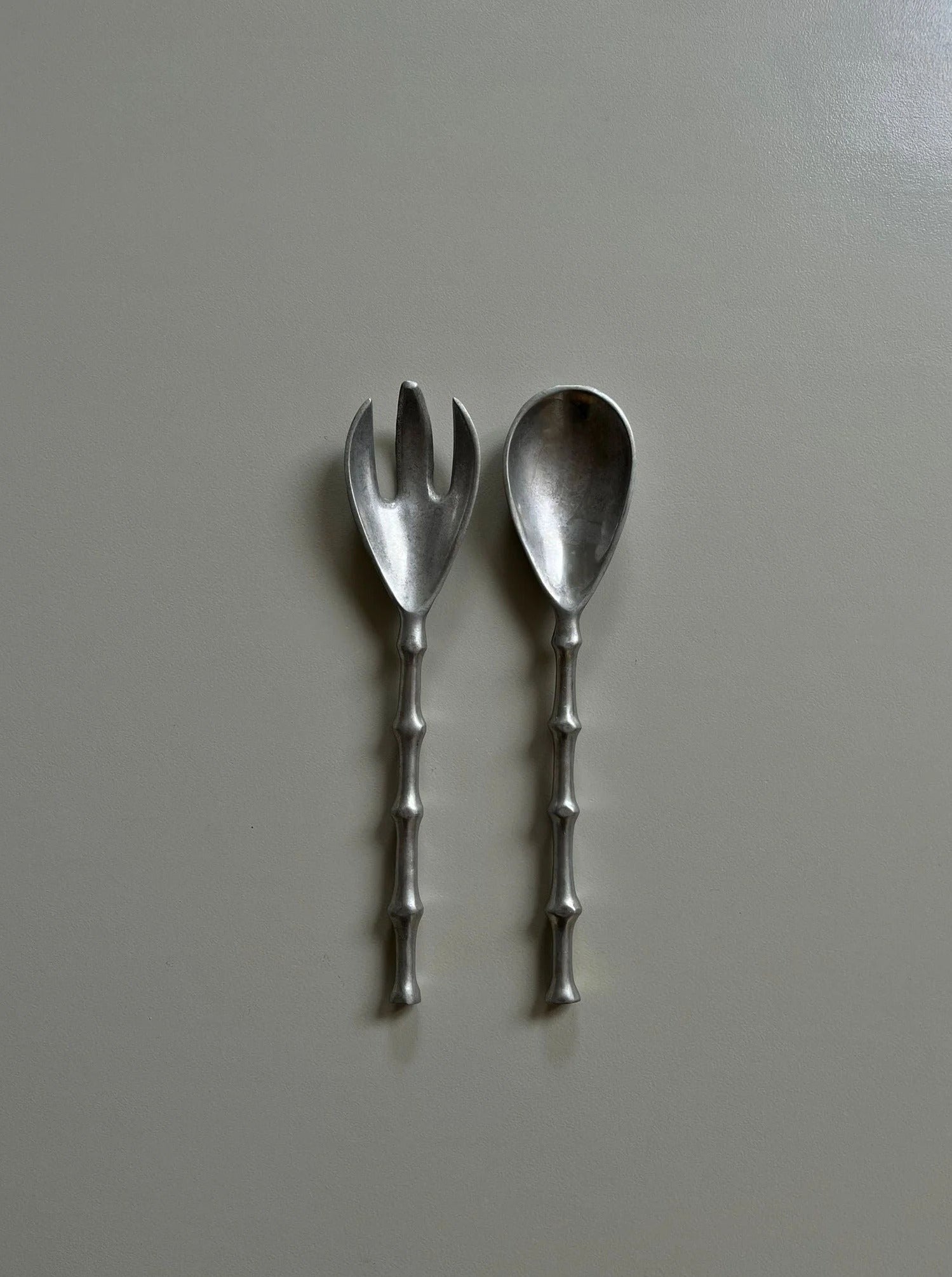 Two silver-plated salad servers, known as Lettuce Cutlery by CU-RA-TED, with handles designed to resemble bamboo stalks are placed side by side on a light gray surface. One serving tool is a fork and the other is a spoon.