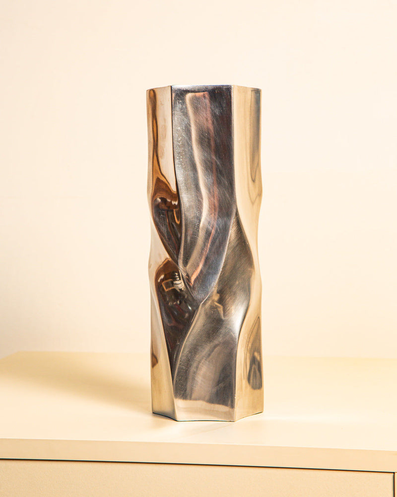 The Treaptyque Large Italian “Hexagon” Vase 80s, a tall metallic cast aluminum piece with a twisted geometric design and shiny surface, stands on a light surface against a pale background.