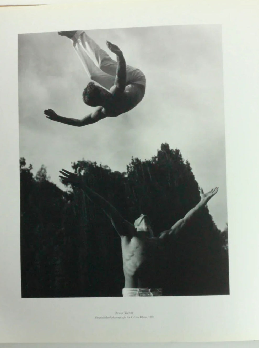 A black and white photograph, part of the exposición en el museo Victoria & Albert Museum, captures a person mid-air during a backflip above another who stands with arms outstretched, ready to catch them. Tall trees frame the background, creating a dynamic and intense scene.