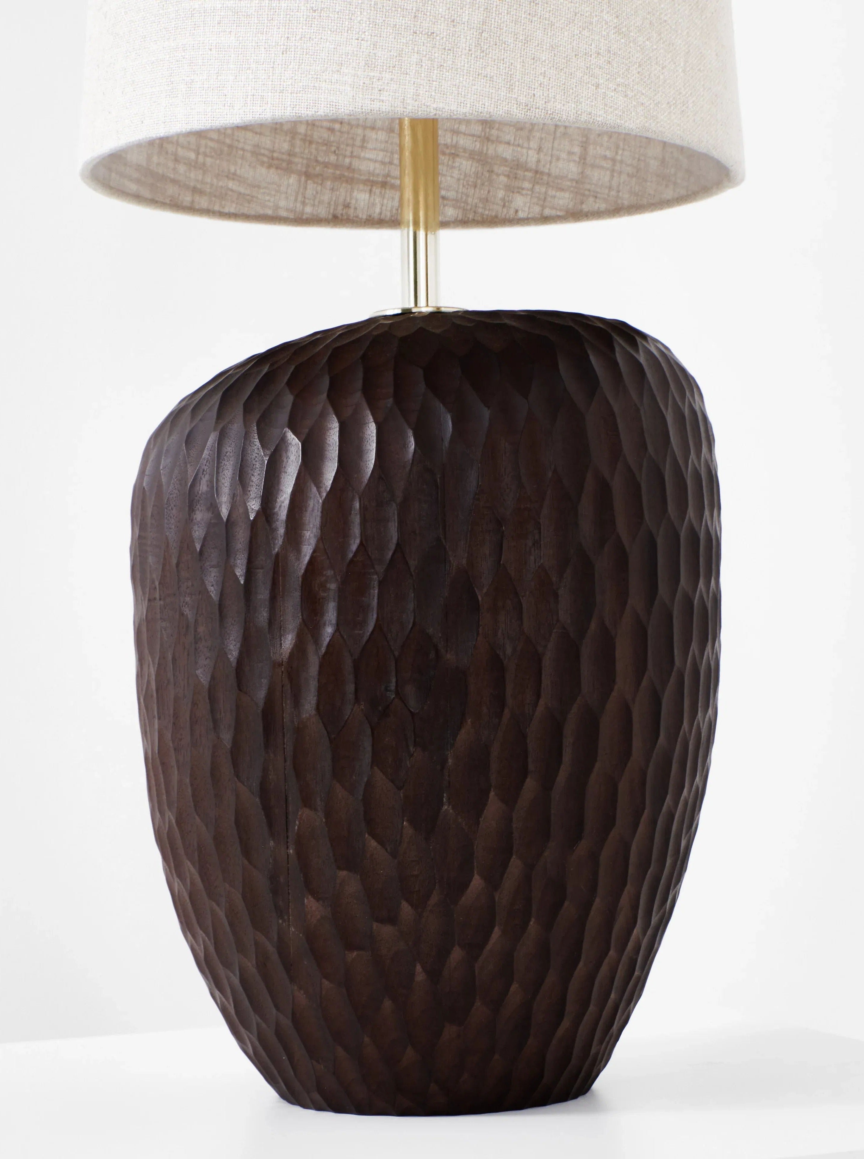 The Large Foz Lamp by Project 213A features a textured, dark brown solid wood base and a beige fabric lampshade. The wooden base has a hexagonal pattern carved into it, adding a rustic aesthetic to the handmade lamp. The cylindrical lampshade has a smooth surface, complementing its artisanal charm.