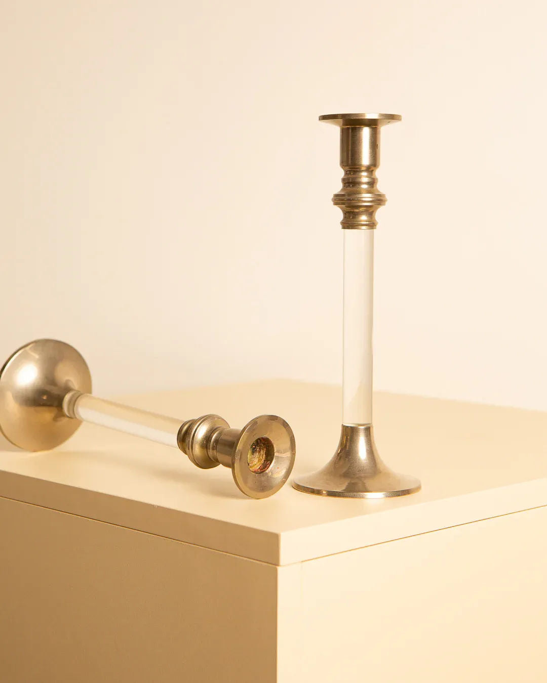 Two Treaptyque Pair of 80's silver and lucite candleholders are placed on a light beige surface. One stands upright, the other lies on its side. The polished finish and slender design exude a subtle elegance, reminiscent of '80s candleholders, blending harmoniously with the neutral background.