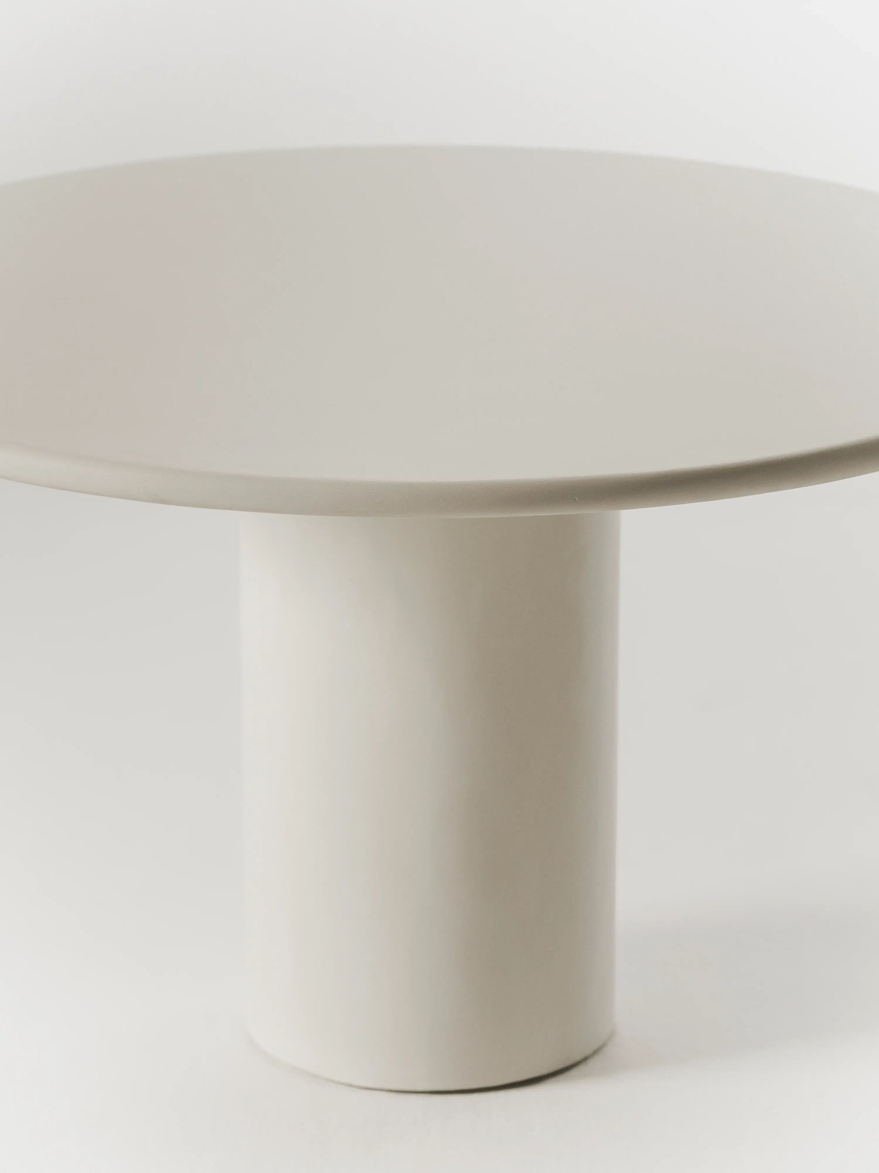 The Gio Dining Table by MarlotBaus features an elegant, minimalist design with a light beige round surface and a cylindrical pedestal base, embodying sophistication against a plain white background.