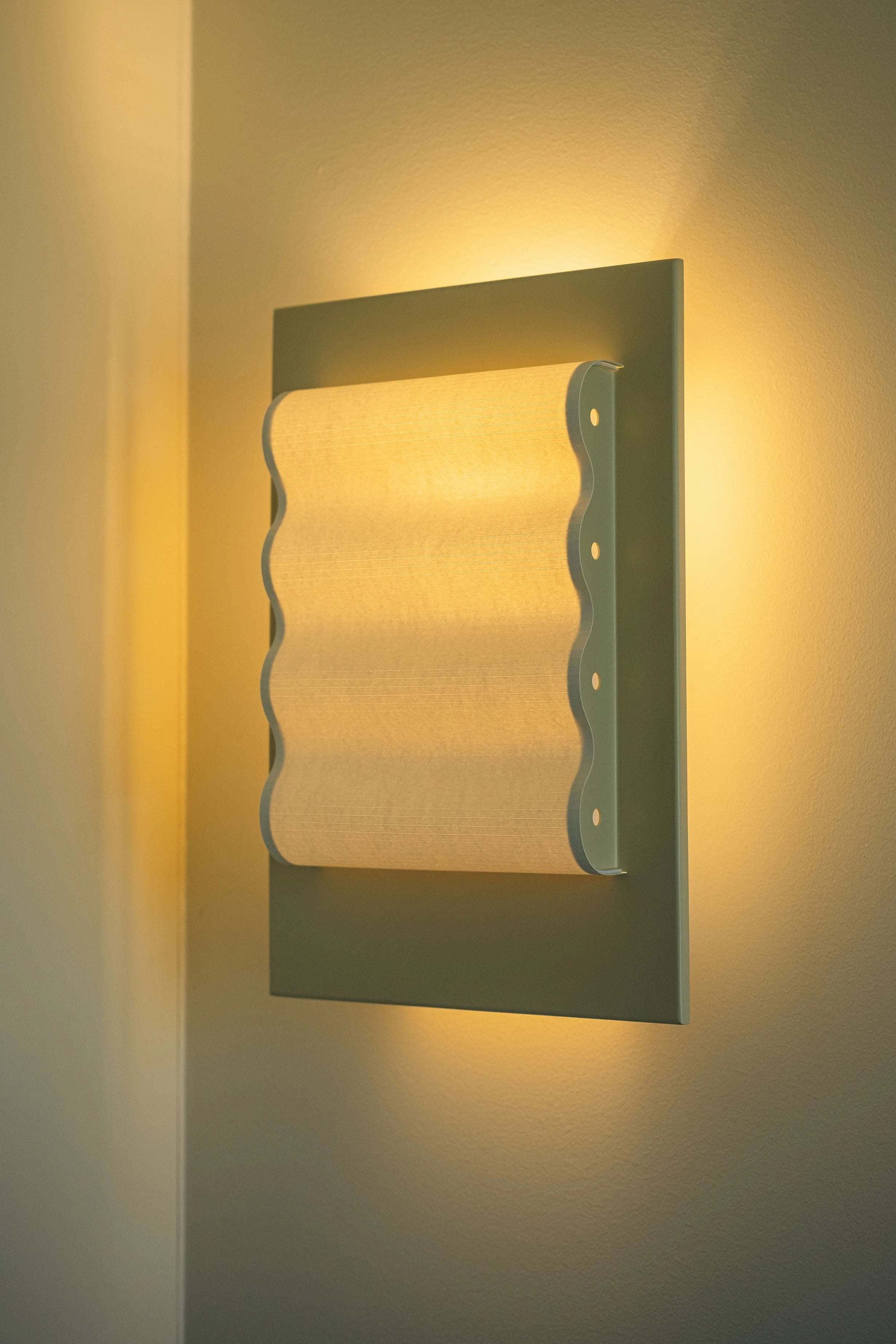 The Violaine d'Harcourt Wall Light Frame OW S features a wavy frosted glass cover that emits a warm yellow light, casting a soft glow on beige walls. This handcrafted, minimalist fixture combines a rectangular frame with smooth curves for modern elegance.