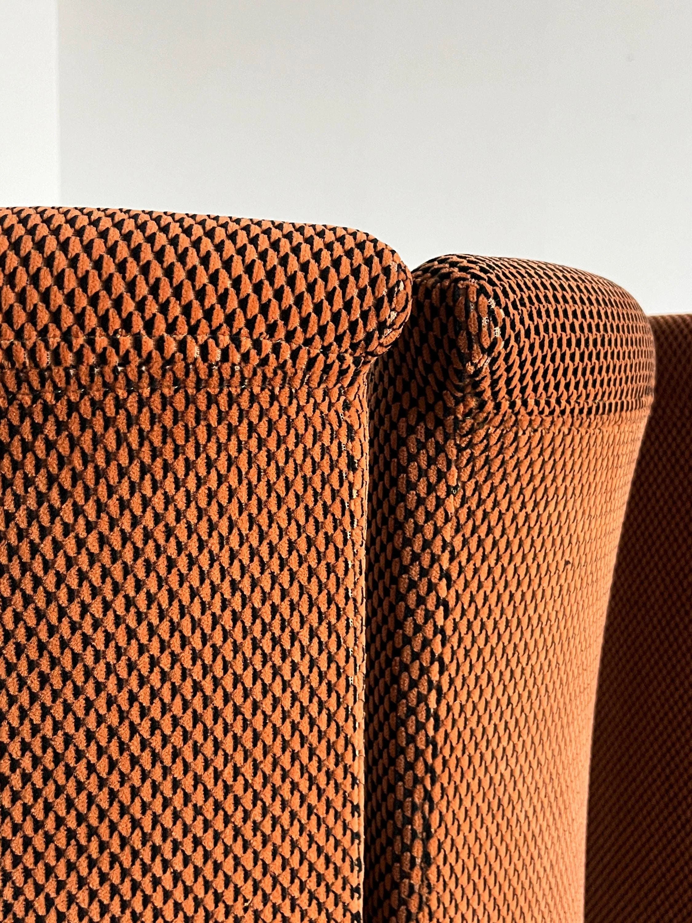 Close-up of Cherry Cargo's Pair of Postmodern Armchairs, featuring orange upholstery with a black geometric pattern, side by side against a white wall. The texture and distinct pattern are clearly visible.