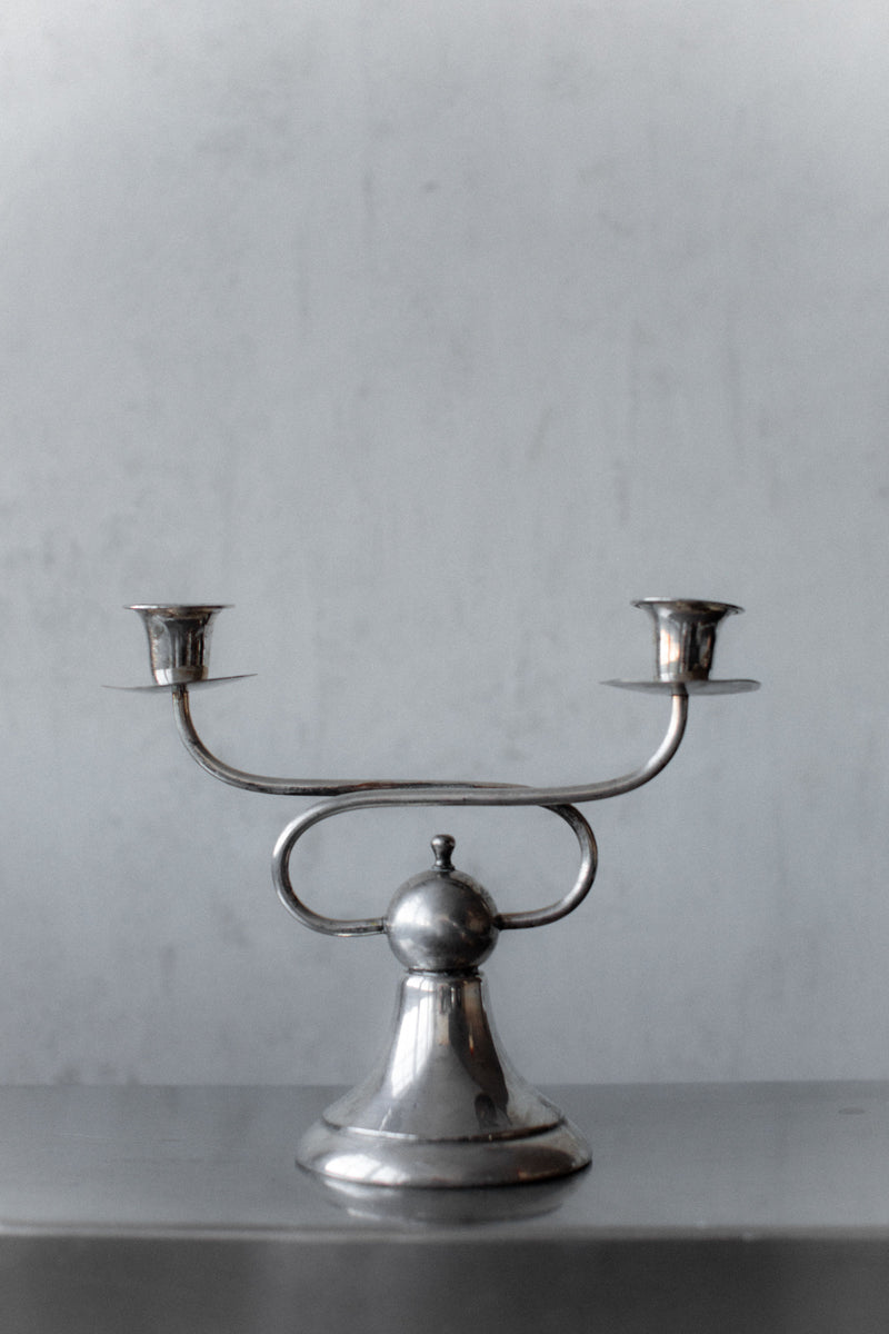 The Art Deco Candleholders by Out For Lunch exude vintage charm with two smooth, reflective arms on a gray background, featuring a central rounded base and elegant curving arms, each ending in a candle socket.
