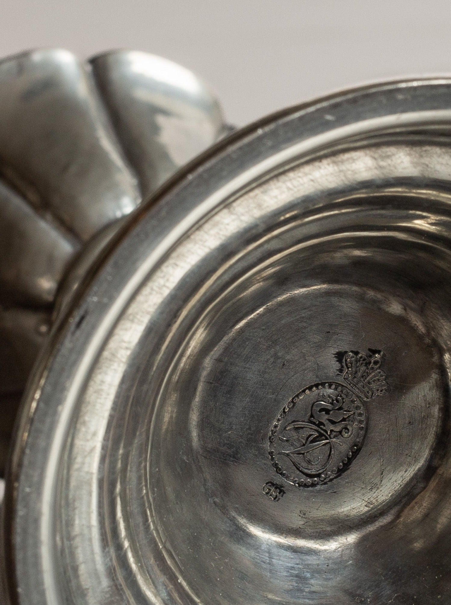 Close-up of the base of a tarnished silver goblet, showing a detailed embossed design with a stylized coat of arms, featuring a crowned emblem at the center with intricate patterns. The background is out of focus, reminiscent of the neoclassical design seen in Spigel's 1930s Art Deco Metal Candlesticks from Denmark.