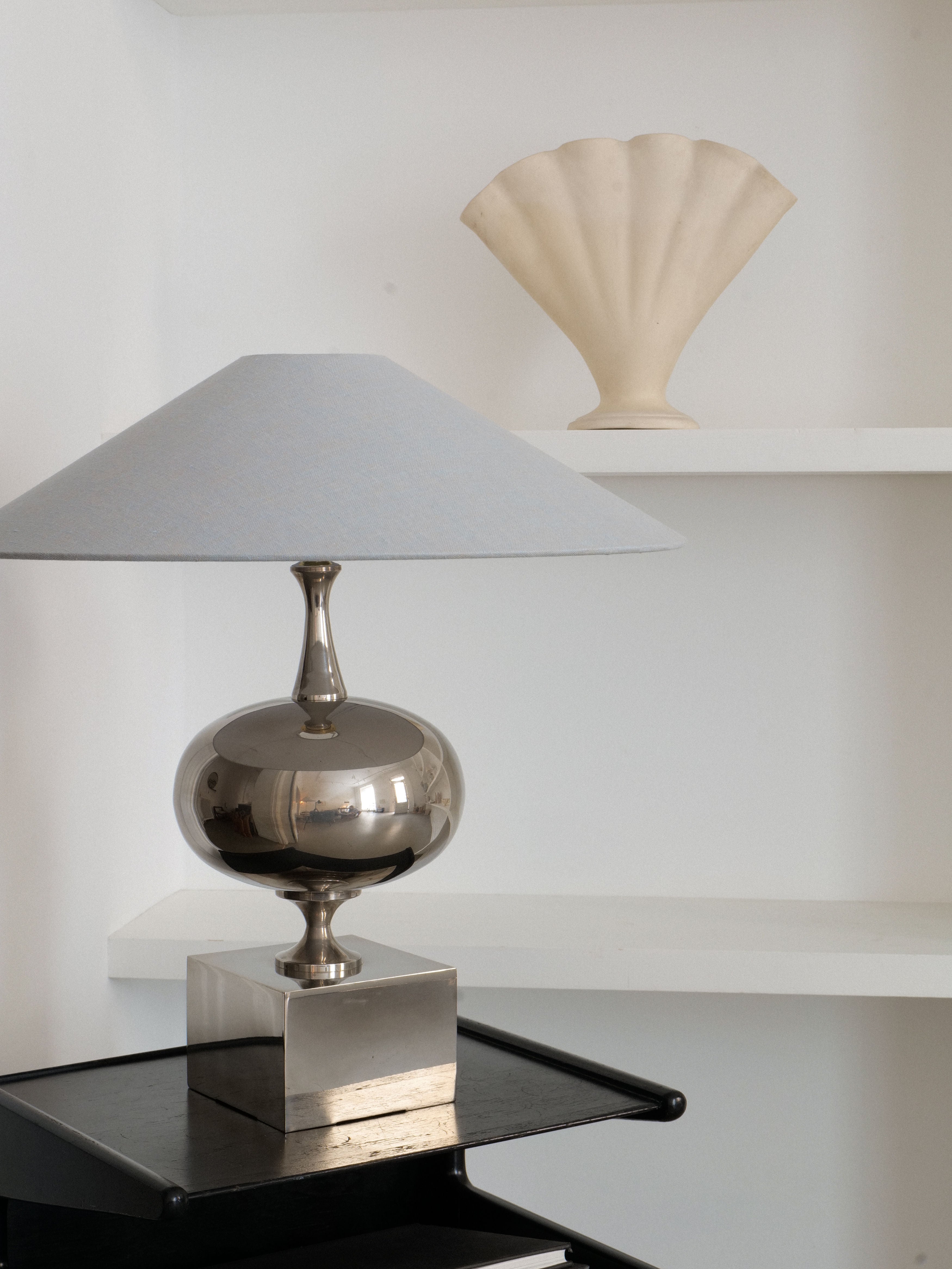 A Philippe Barbier Sculptural Lamp from the 1970s by Collection Apart, featuring a chrome over brass base and a blue shade, sits on a black side table in front of white shelves. On the upper shelf, there is a beige seashell-shaped decor piece. The background is minimalistic, with a neutral color palette.