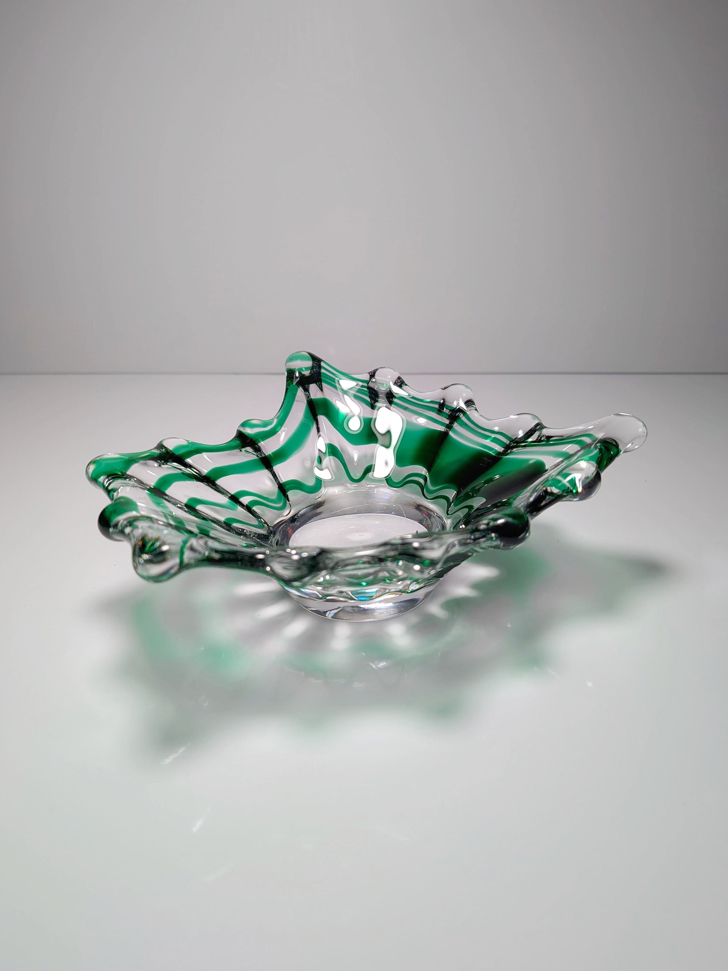 The Green Swirled Murano Vide-Poche by Dodo Vintage, a transparent glass bowl with a wavy, decorative rim and green-clear swirl pattern, sits elegantly on a reflective surface against a neutral background.