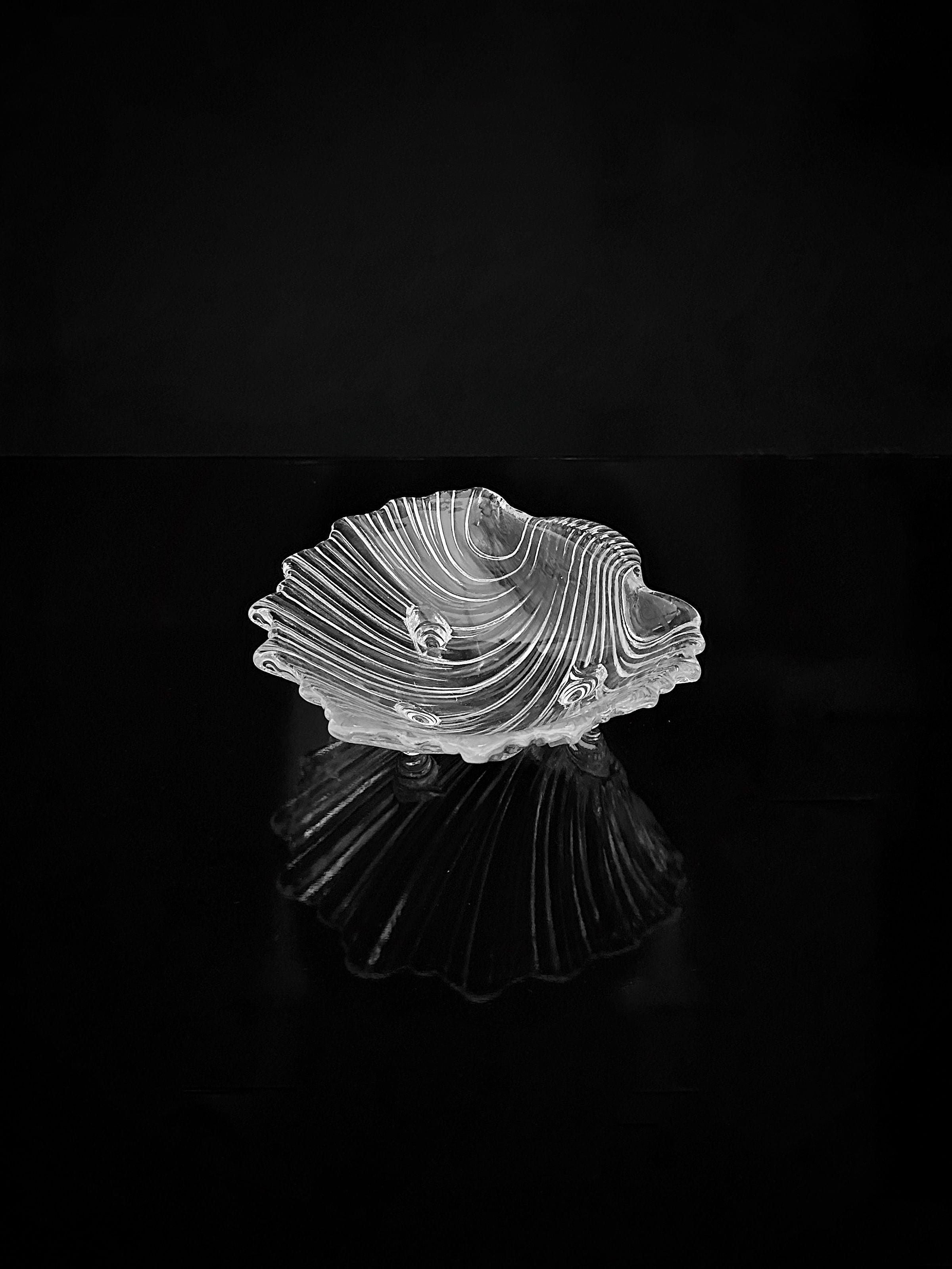 A 1980s Shell Shaped Glass Bowl by Dodo Vintage, artfully designed with wavy ridges and layered textures, rests on a glossy black surface, creating a subtle reflection. Its elegant shape is perfect for the sophisticated presentation of delicacies.