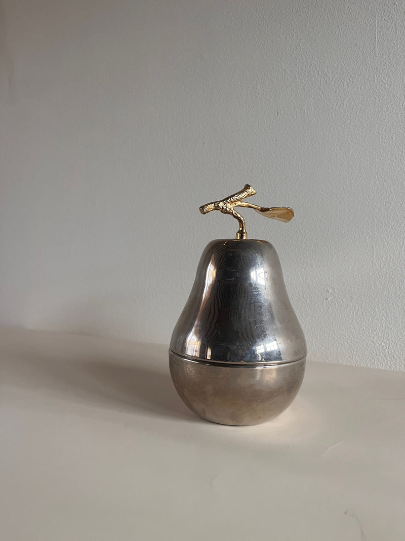 The Maud Vaughan Mid Century Pear Lidded Box, crafted from mixed metal with a golden stem and leaf, sits on a light surface. Its shiny texture and elegant design give it a modern mid-century artistic appearance.