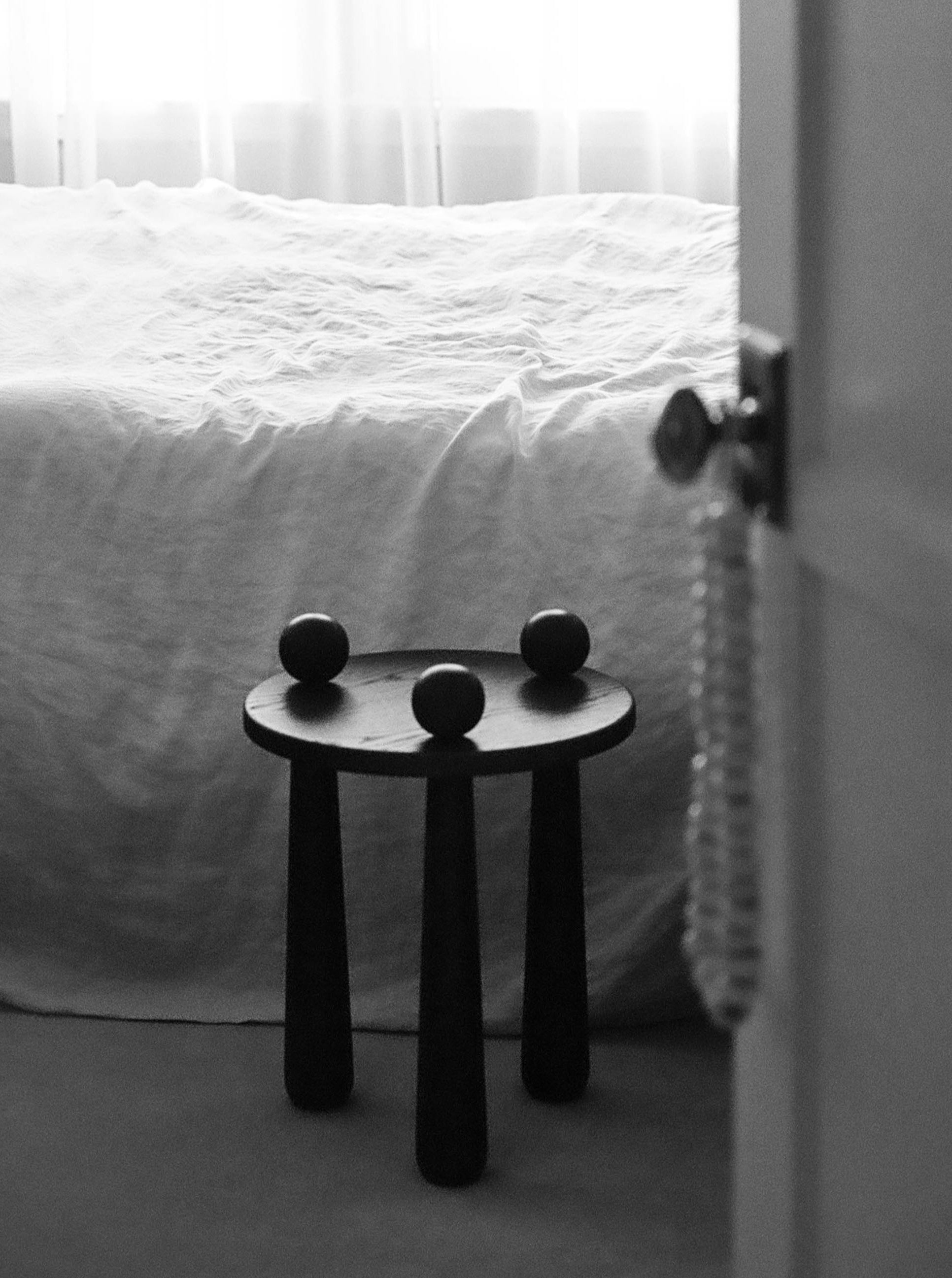 Black and white image of a minimalist interior. A small, round, three-legged Dott Stool, Ebony by TASE GALLERY sits next to a bed covered with a plain, white sheet. Three small, round objects like ball and bass caps are placed on the table. Part of a door and a doorknob with a beaded decoration are visible.