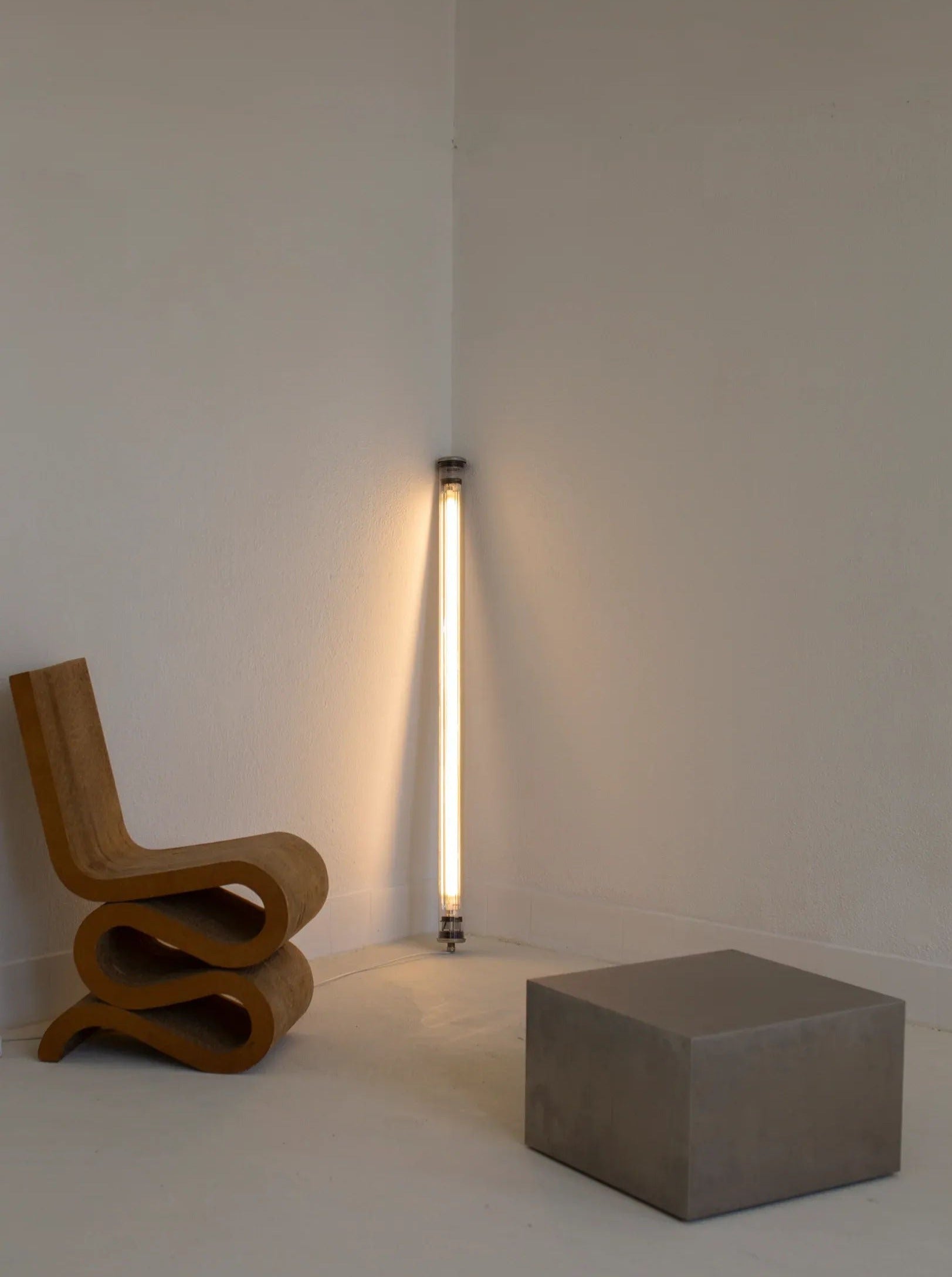 A minimalist room features a sculptural wooden chair to the left, a CU-RA-TED Sammode Light Tube leaning against the corner of the walls, and a small, square concrete block as a low table or seat in the foreground. The room's white walls and floor highlight its understated elegance.