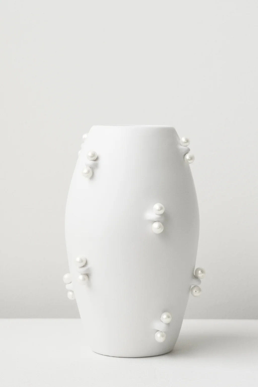 The "Pinched" small vase in matte white by Completedworks features a sculptural form with clusters of raised, pearl-like accents, displayed on a light-colored surface against an off-white background, blending traditional ceramics with modern design.