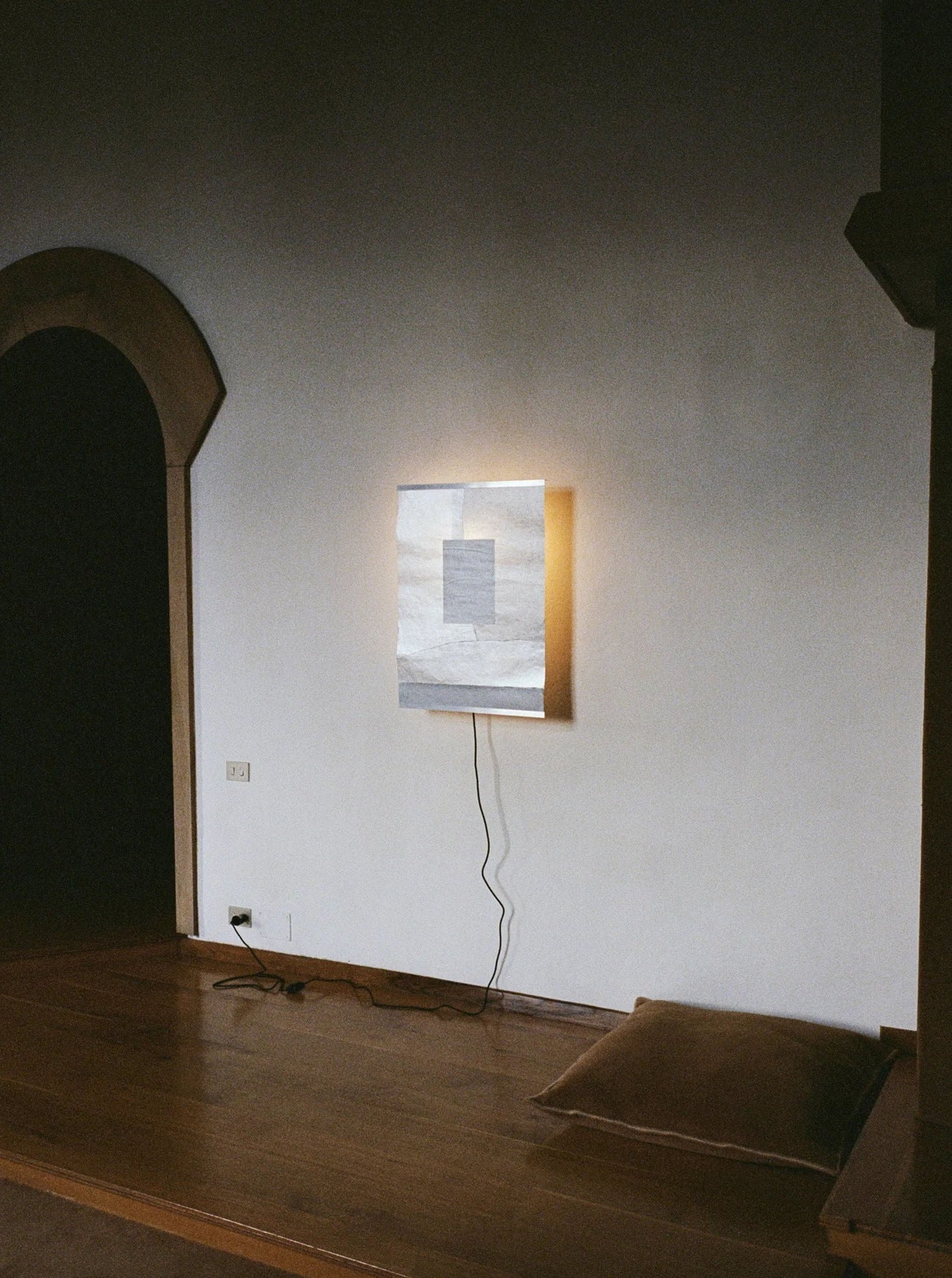 In a minimalist room, a softly glowing TEGET Panel Wall Lamp – "Icescape" is mounted on the wall. A pillow rests on the wooden floor, and a dark archway is visible to the left. The lamp's black cord trails across the floor.