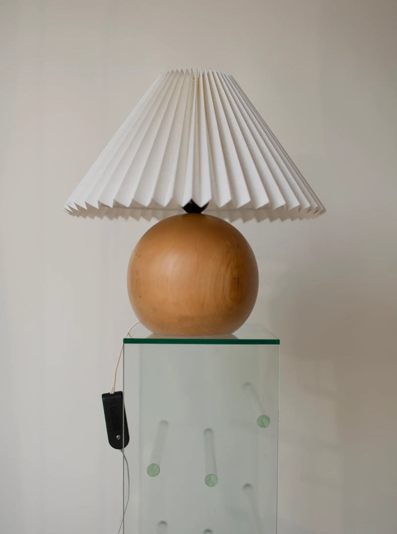 A unique CU-RA-TED Wooden Ball Lamp With Plisse Shade offers an elegant lighting fixture. Placed on a white surface, it complements the black smartphone leaning against the base, connected to a white charging cable. The use of natural materials gives it a timeless appeal against the plain white background.