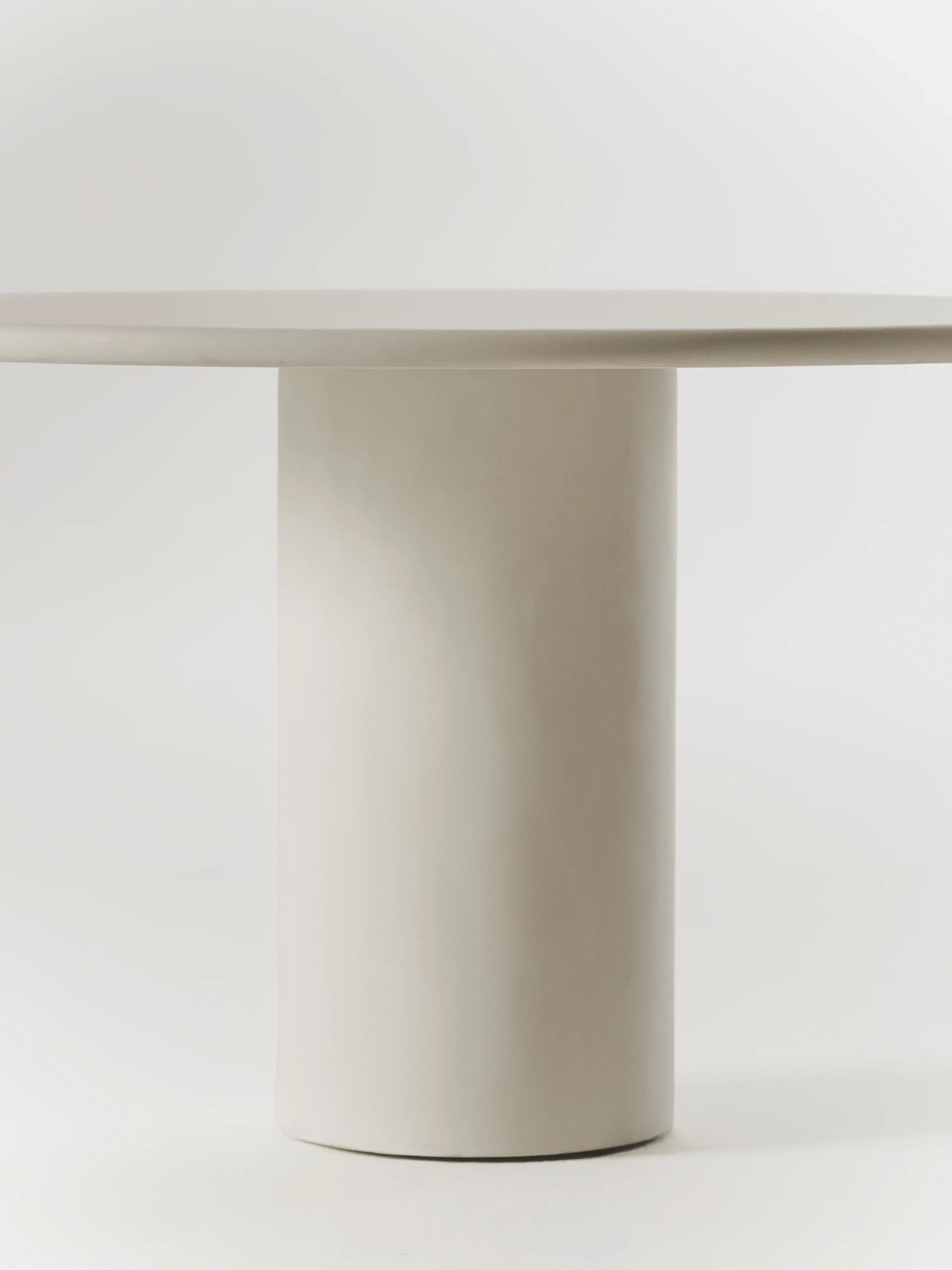 The Gio Dining Table by MarlotBaus is a minimalist microcement piece with a cylindrical base and round flat top in a neutral light color, elegantly set against a plain background.