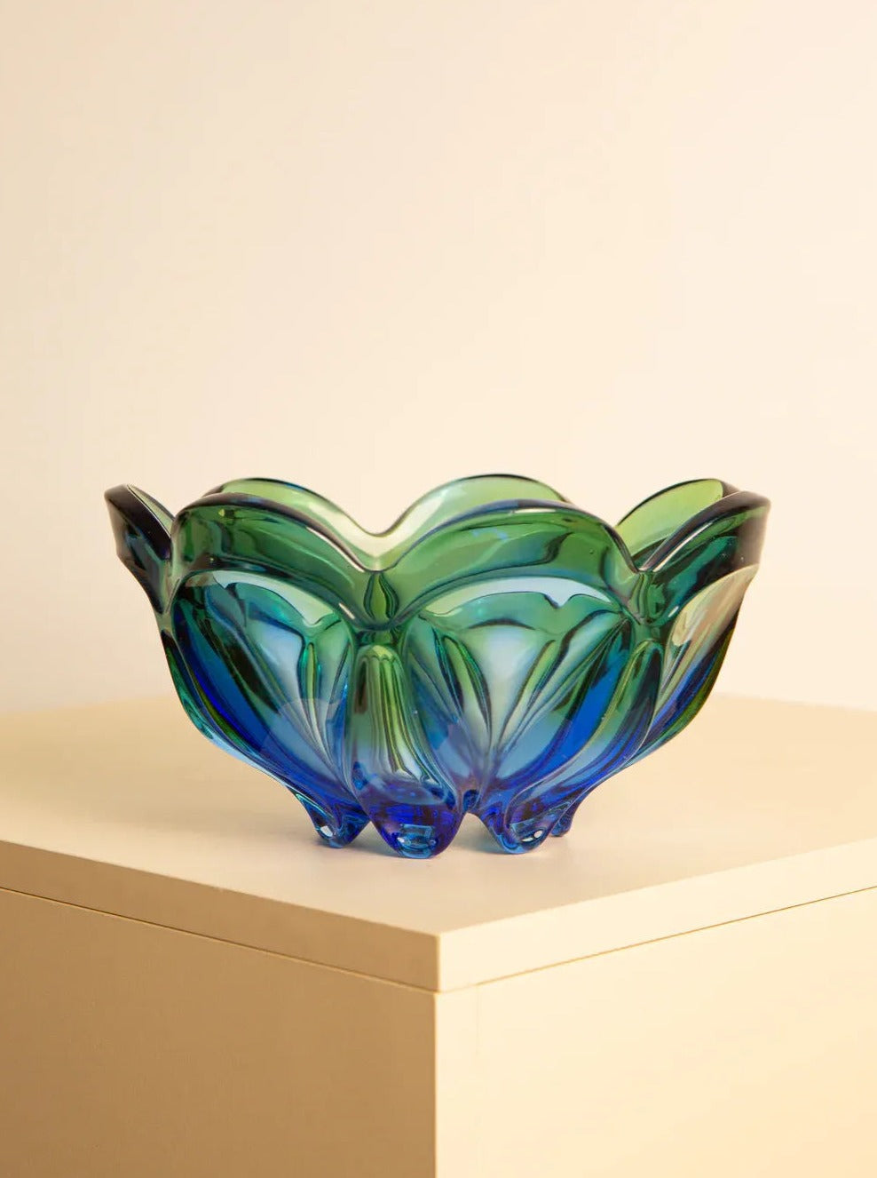 A Large vide-poches "Flower" in 60's Murano glass by Treaptyque with a wavy, scalloped edge in shades of blue and green sits on a light-colored wooden surface. Reminiscent of 1960s retro design, the bowl’s multiple folds and curves give it a textured and artistic appearance. The background is plain and softly lit.