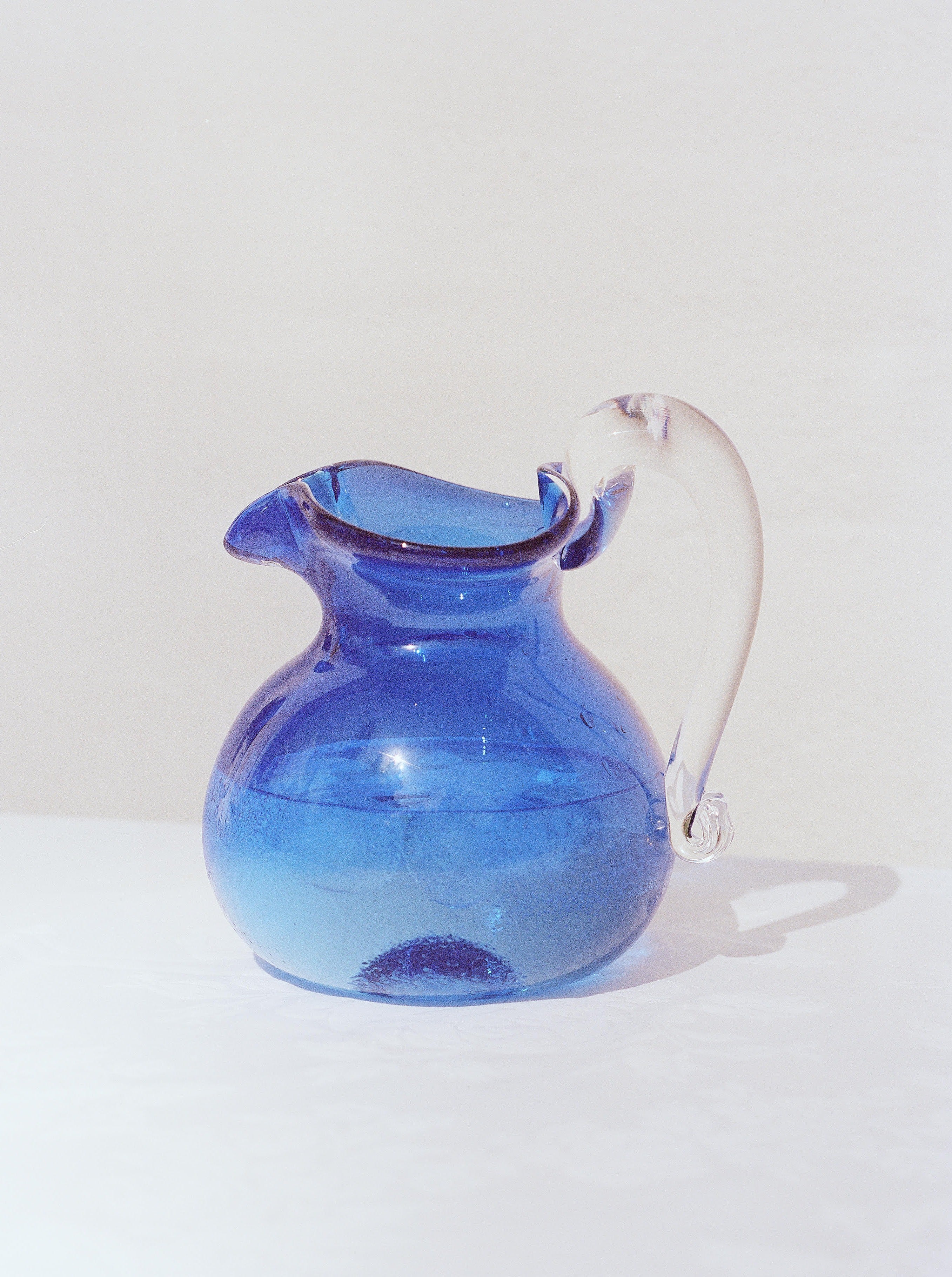 A blue Michele Jug from Akua Objects with a rounded body and a clear, curved handle sits on a white surface against a plain beige background. The handmade glassware boasts a smooth texture and reflective quality, while the handle features a decorative flourish at the top.