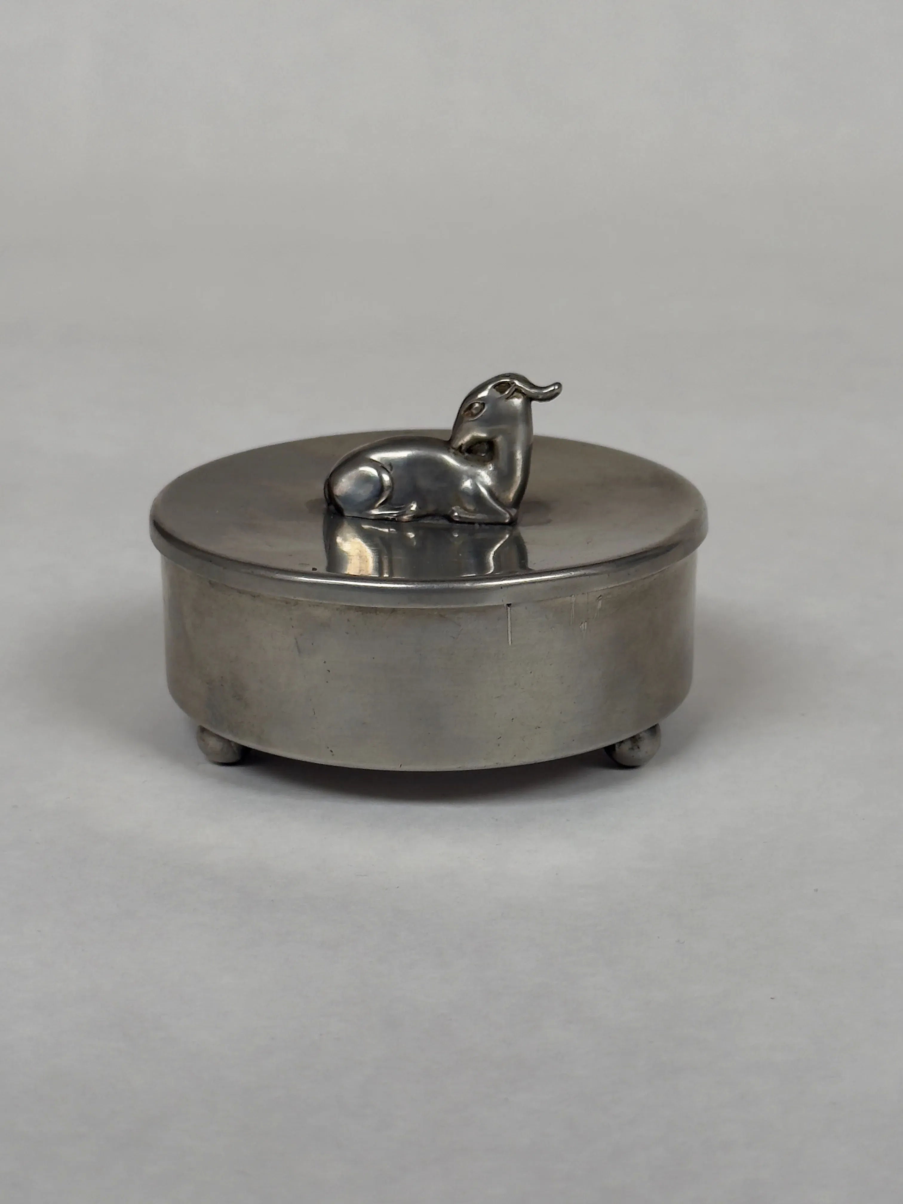 The Galerie Storm Vintage Pewter Trinket Box is a round silver treasure with a lid, featuring a sculpted elephant handle. It rests on four spherical feet, ideal for storing jewelry against a light gray backdrop.