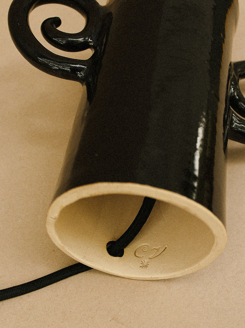 A close-up image of a ceramic object with a shiny black glazed exterior and an unglazed interior. The hand-built stoneware Espiral Lamp - Grande black by Casa Veronica features curly handles and a hole at the bottom where a black string is threaded through. A symbol is impressed on the inner, unglazed surface.