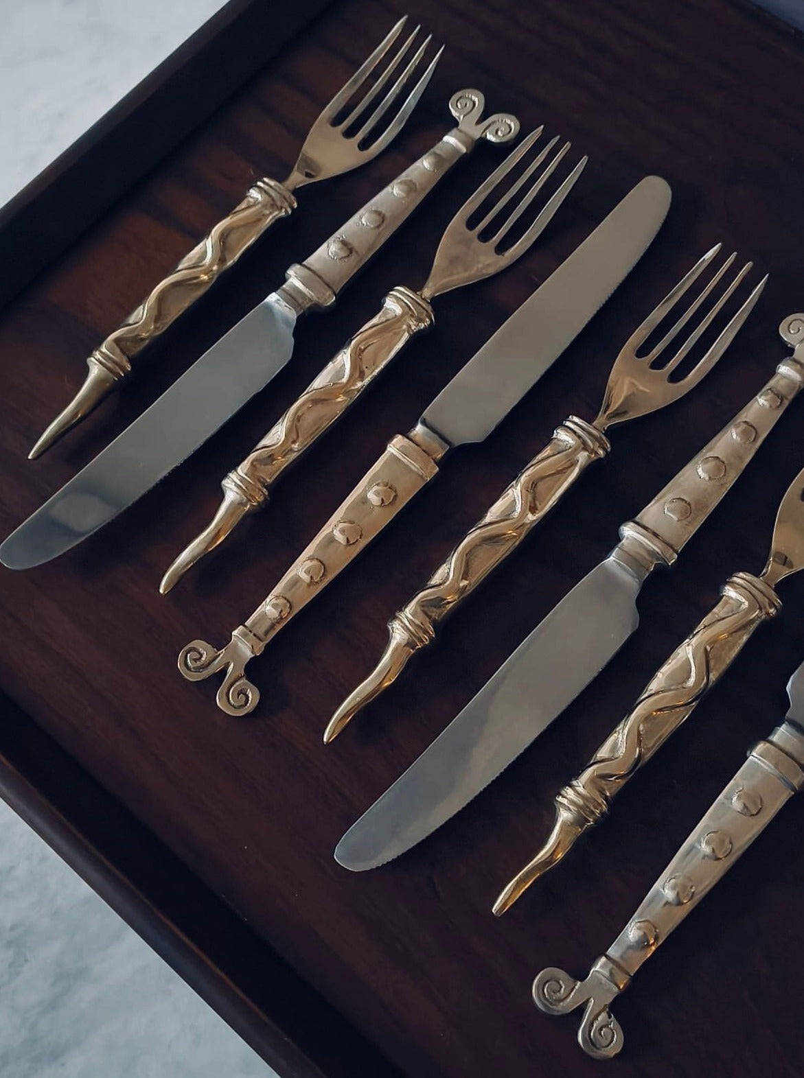 The Médecine Rare Vintage Cutlery Set is housed in a wooden display case and includes four knives and four forks, neatly arranged alternately. Each piece features stainless steel blades paired with ornate, intricately designed handles adorned with elegant curved patterns in gold and silver tones.