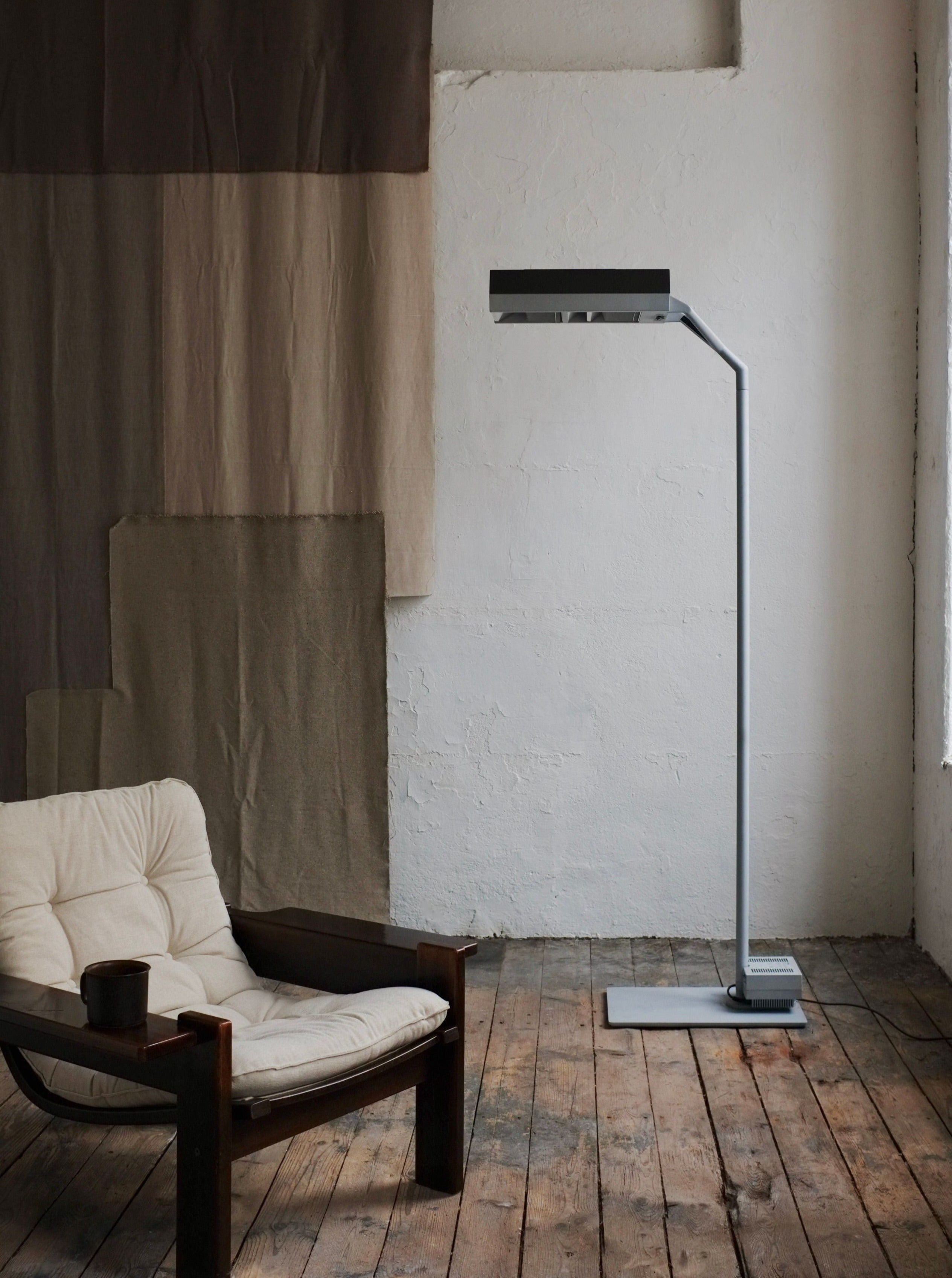 A minimalist corner with an armchair and a Postmodern Metal Lamp by Glen Oliver Löw and Antonio Citterio for Ansorg | 1990s beside it, set against a rustic white brick wall with a large draped fabric piece. A wooden floor and a small cup on the armrest add cozy details.