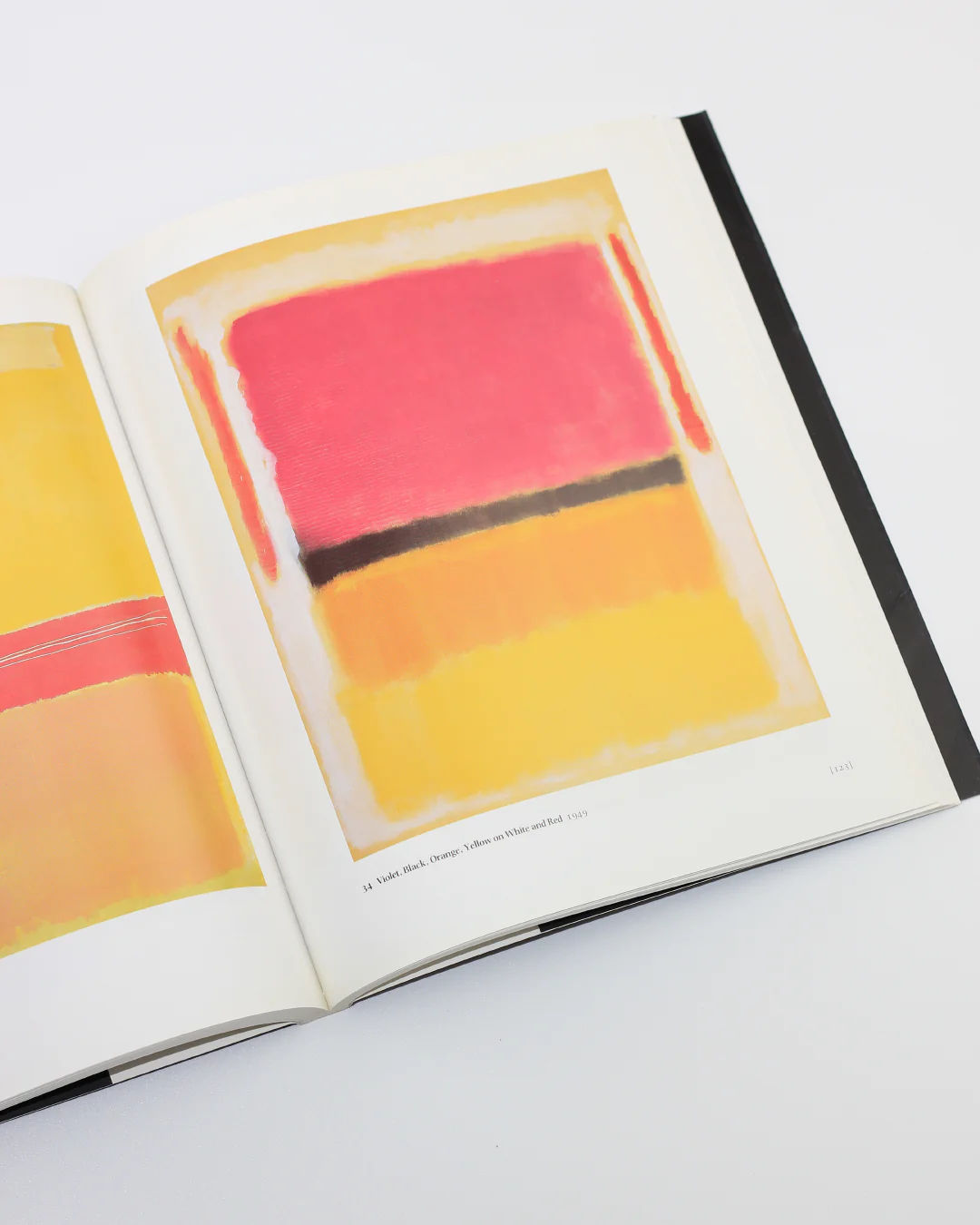 The Mark Rothko Book by Boga Avante Shop lies open, showcasing a painting with horizontal color blocks in pink, black, and yellow. A page featuring Abstract Expressionism is partially visible on the left as the book rests on a white surface.