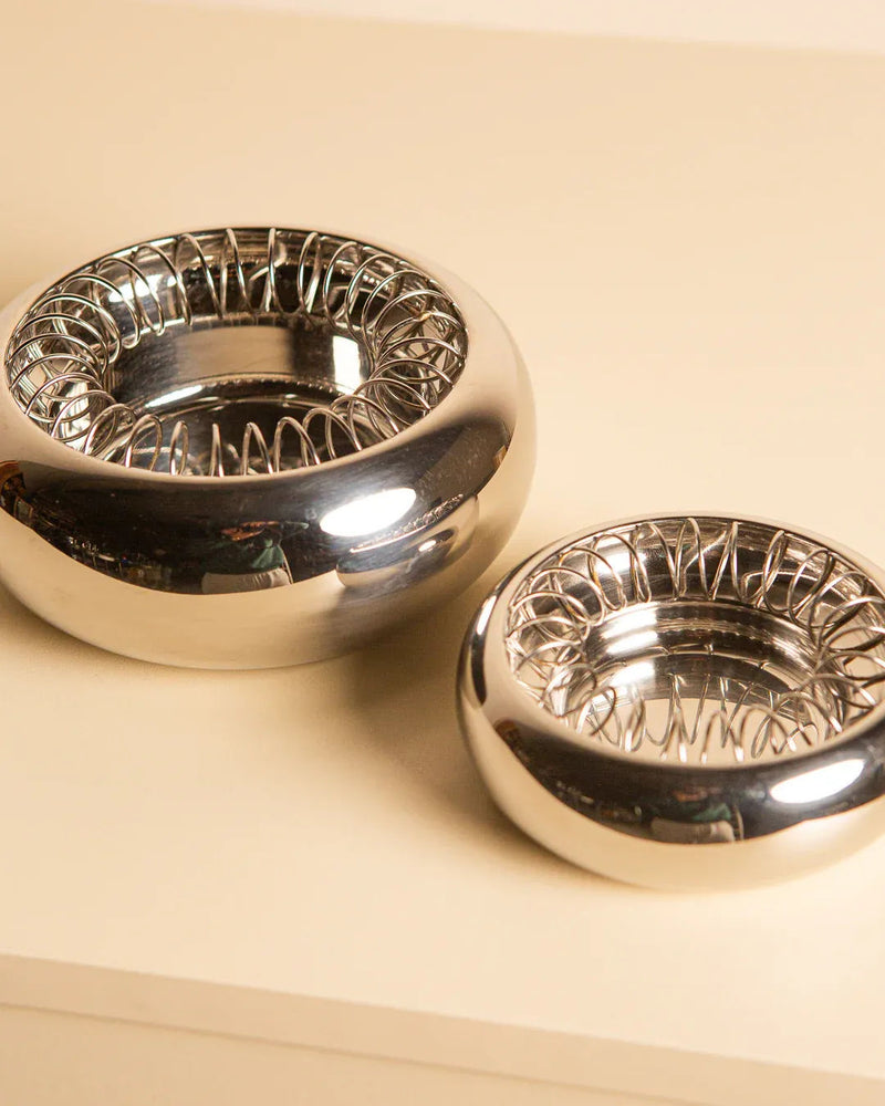 A pair of "Spirale" ashtrays by Achille Castiglioni for Alessi 90s, from Treaptyque, rests on a beige surface. With a shiny, spiral design and smooth finish, one larger than the other, they subtly capture and reflect light in true Italian style.
