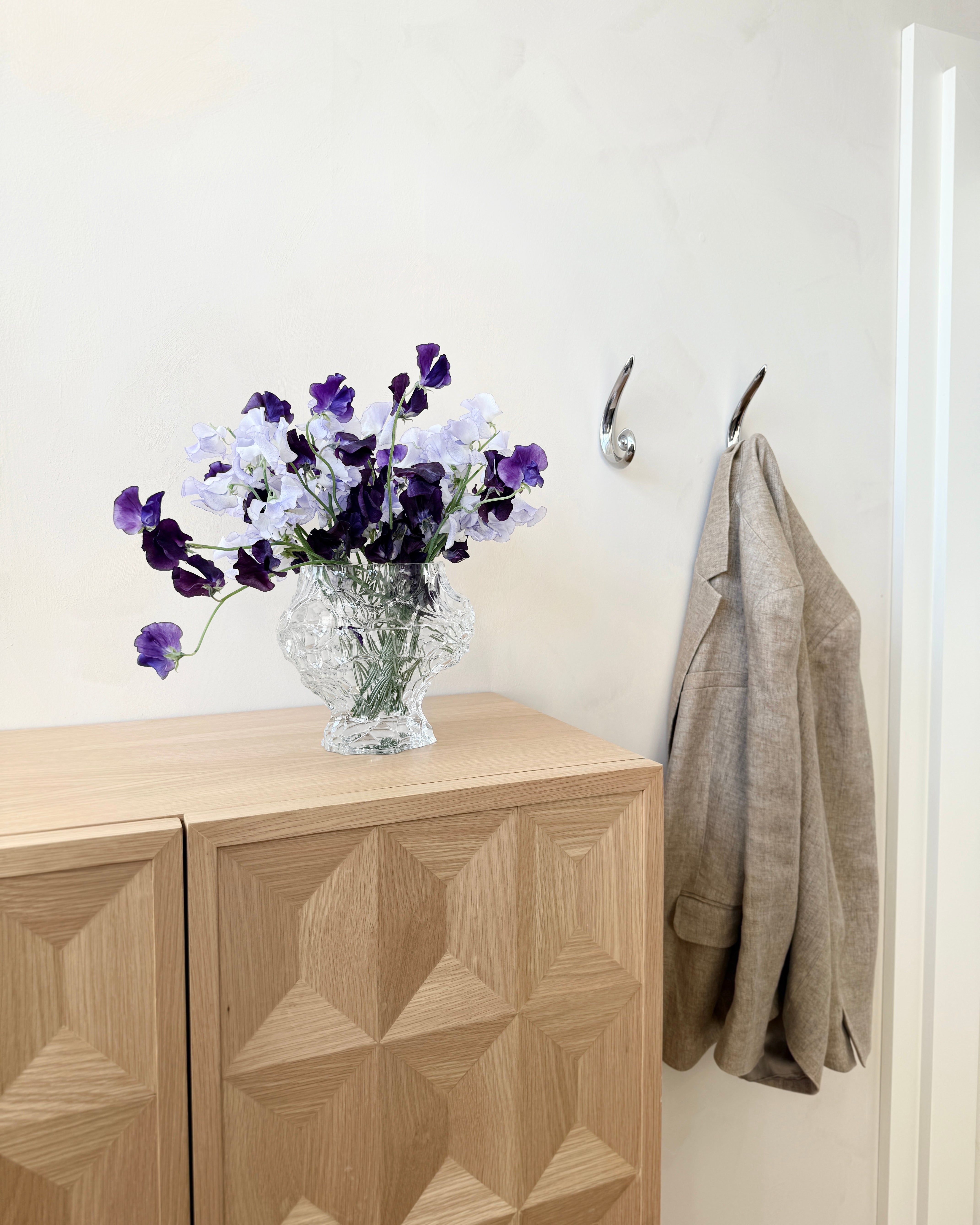 A glass vase with purple and white flowers rests on a wooden cabinet. Nearby, a light gray blazer hangs on Hein Studio’s Noelle Hook in brass, adding elegance against a light-colored wall.