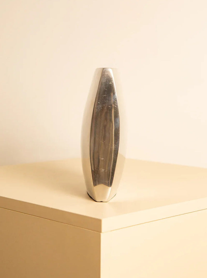 A sleek, Treaptyque 80's Italian metal vase with a modern, elongated shape stands on a beige minimalist pedestal against a light beige background. The vase's silver metallic and reflective surface subtly mirrors the surrounding light.