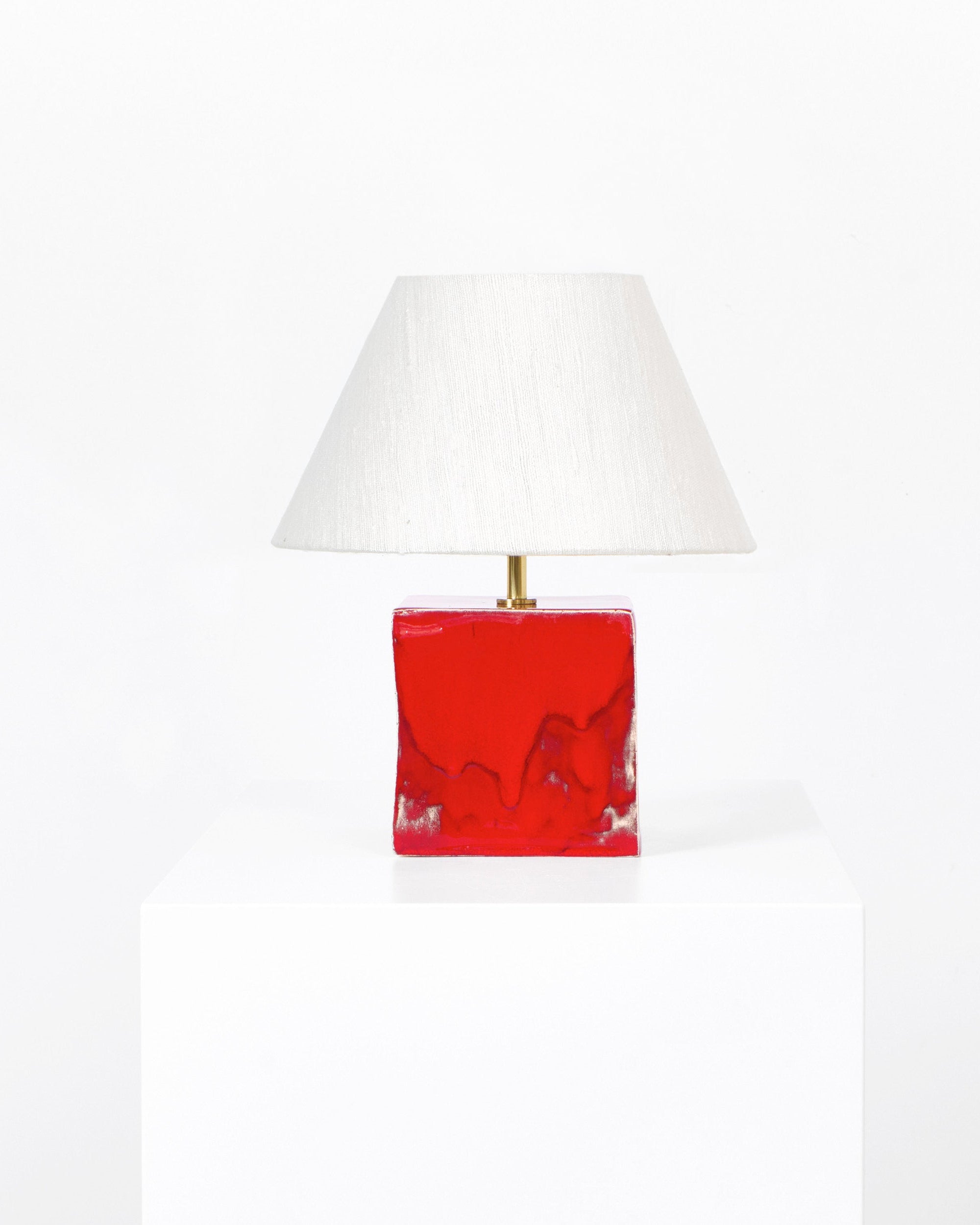 The Arouca Table Light Red by Project 213A features a glossy red, rectangular ceramic base and a white, tapered lampshade, displayed on a white pedestal against a plain background to highlight its handmade elegance.