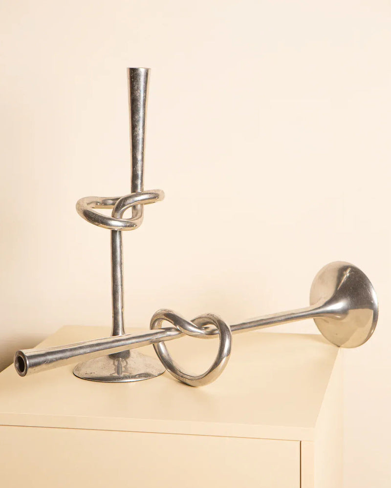 Two Treaptyque "Nœud" aluminum candleholders from the 80s, with twisted knot designs, rest on a light surface; one stands upright while the other lies horizontally, highlighting their unique sculptural forms against a neutral background.