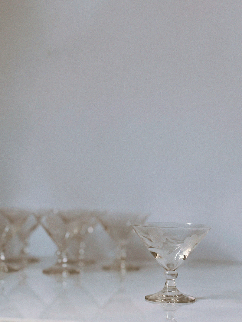 Eight floral etched glasses by Huerto are arranged on a white surface; seven form a group in the background while one is in clear focus at the front, set against a plain light backdrop.