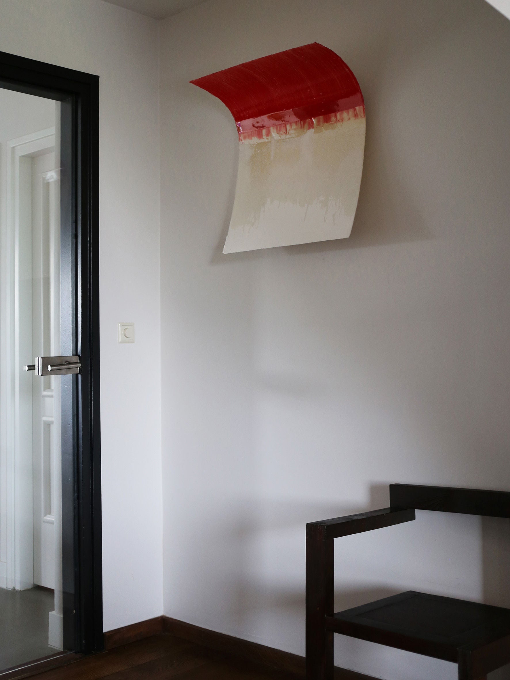 In a minimalist room corner, the serene atmosphere is completed by STUDIOPOLS's "Red Curve" Epoxy Sculpture—a unique piece of modern art featuring a bent, rectangular sheet painted red on top that transitions to white. A dark wooden chair and a black-framed glass door enhance the space.