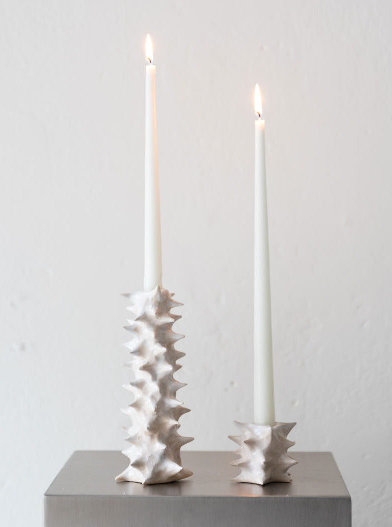 Two white taper candles are lit and placed in unique 2222Studio Spiky ceramic candle holders of different heights, set on a metallic surface against a plain white background.