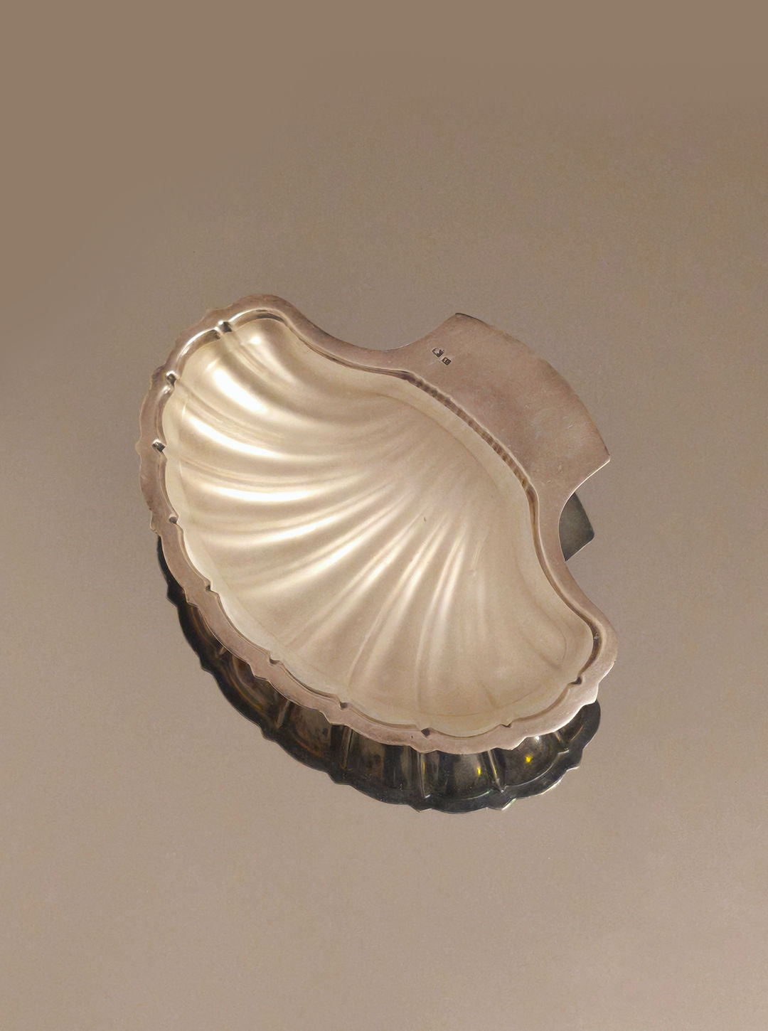 The Shell Butter Dish by Les Objoies is a silver-plated, shell-shaped serving piece. It features a smooth, polished surface with curved ridges reflecting a seashell design on a plain background.