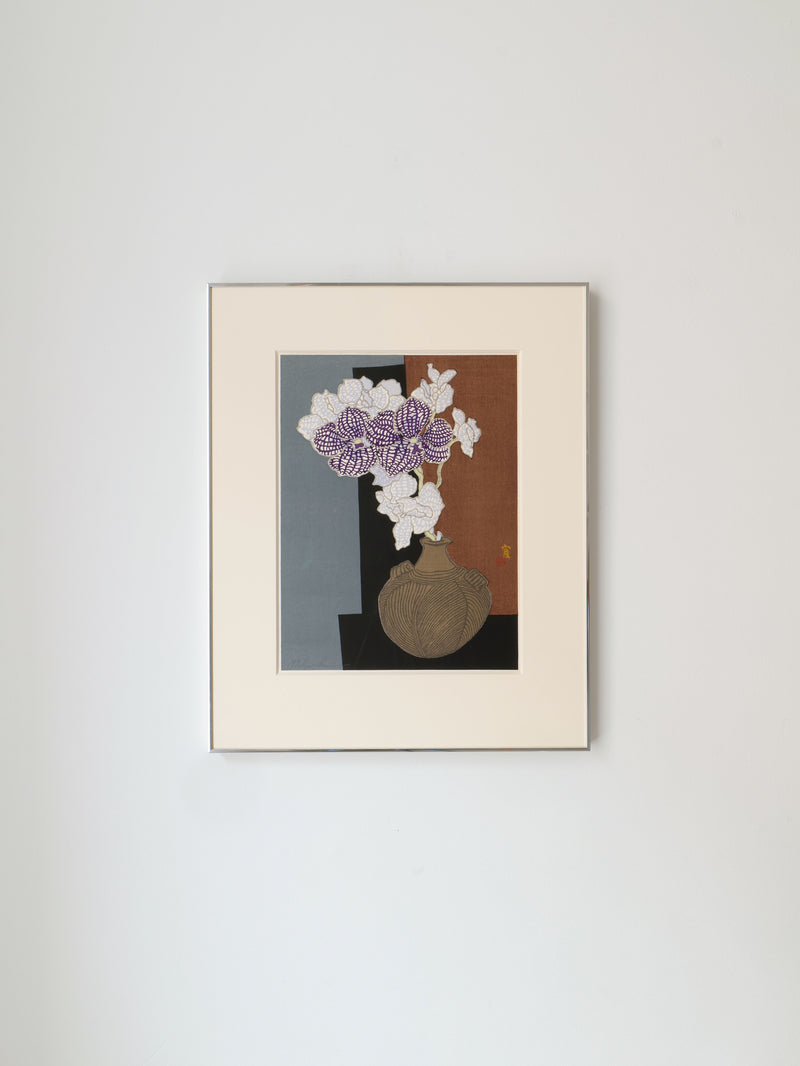 An off-white framed artwork resembling Tokuriki Tomikichiro's Japanese woodcut print from the 1980s showcases a vase with white and light purple flowers on a muted gray-brown background, evoking the delicate beauty of an orchid. It is part of Collection apart.