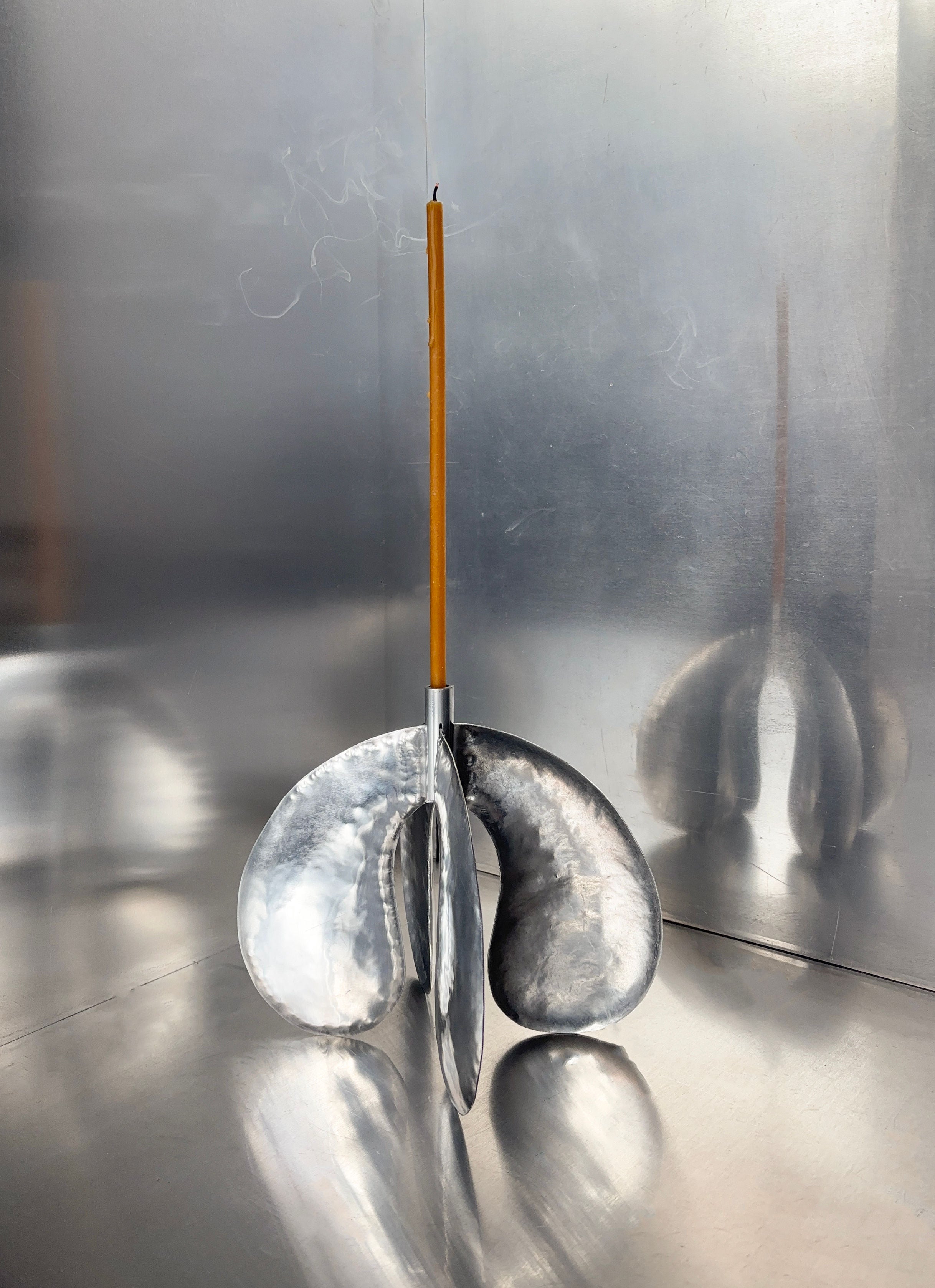 A sleek, hand-made Phaedra candle holder LARGE by Bregje Sliepenbeek features an abstract, curved metal base on a reflective surface. A lit candle is centered, with smoke gently rising above, embodying Amsterdam's polished and modern craftsmanship.